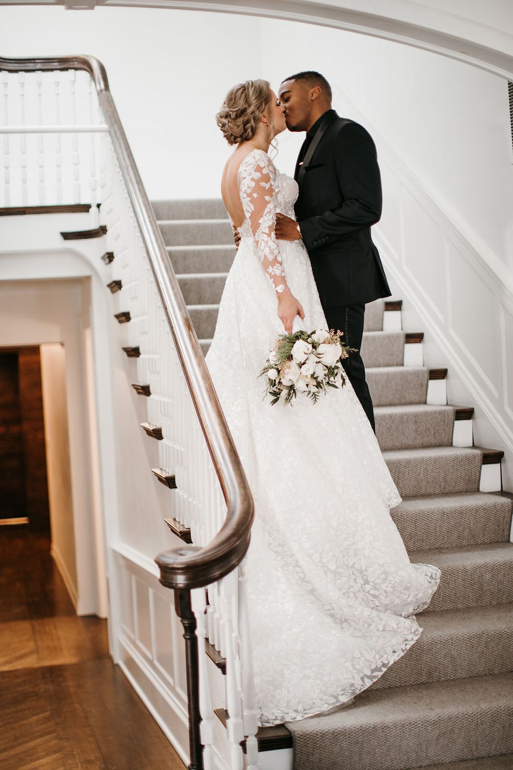 Bride and groom sharing a kiss on elegant staircase, bride in lace gown with bouquet.