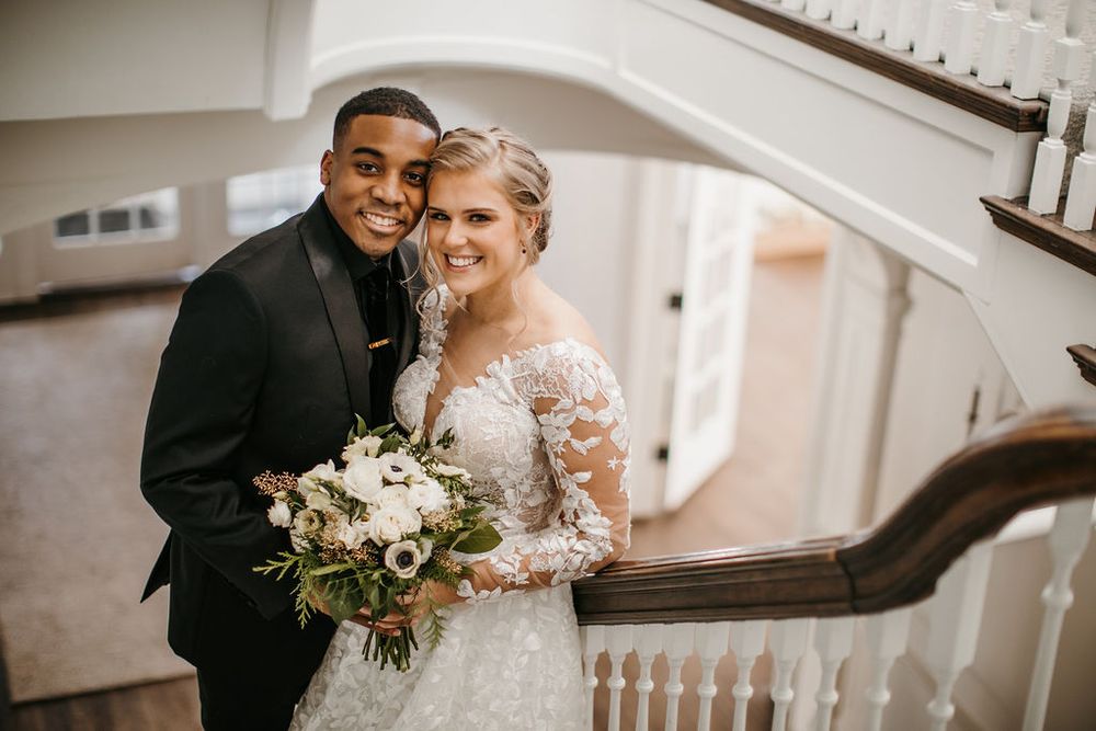 Interracial wedding couple smiling on grand staircase, bride holding white and green bouquet.