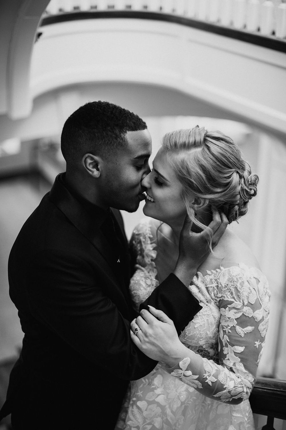 Black and white photo of interracial bride and groom sharing romantic kiss on staircase.