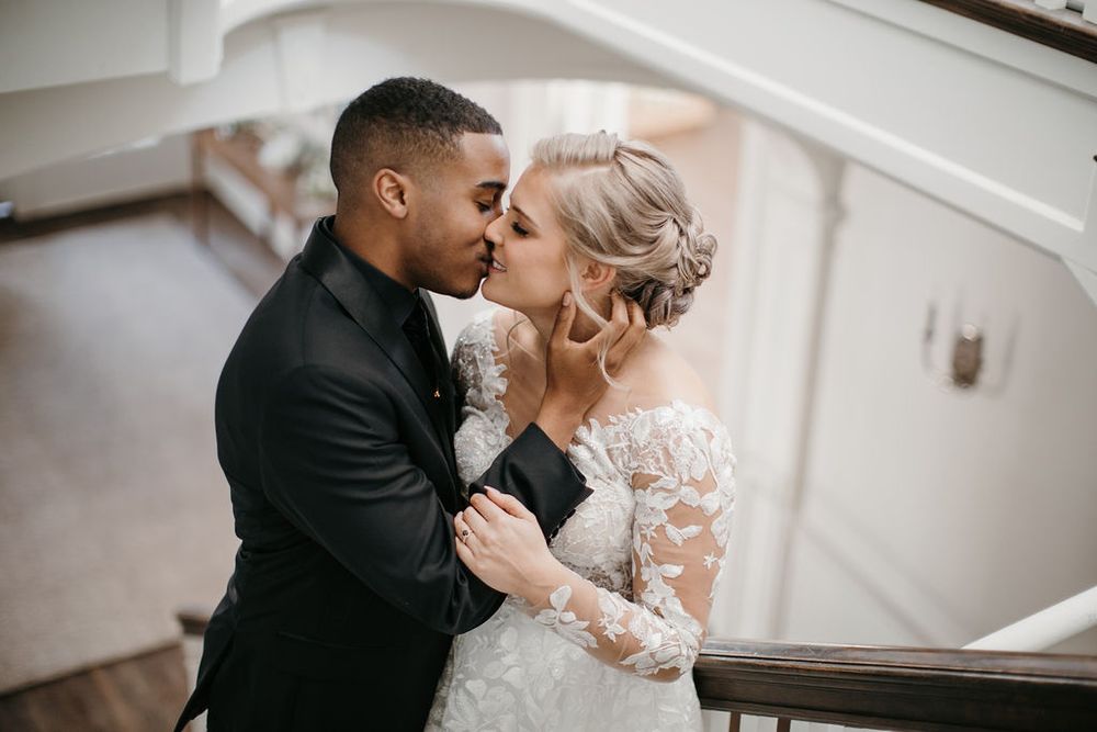Interracial couple sharing a romantic kiss on a staircase, bride in lace gown and groom in black suit.