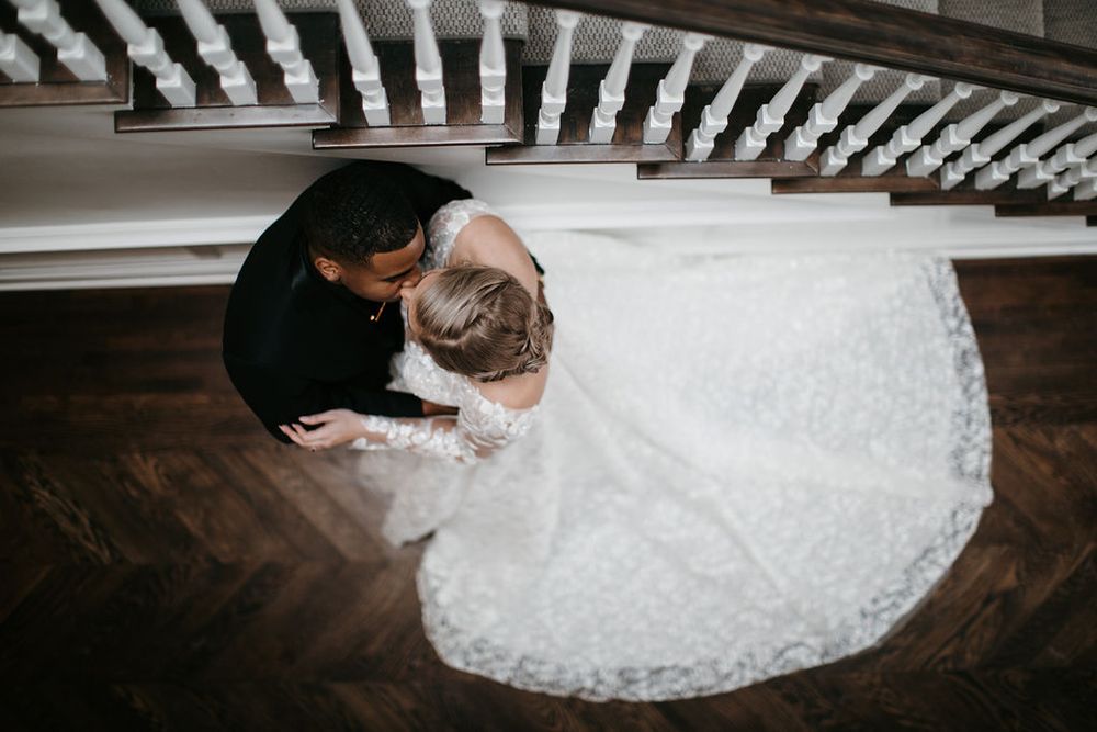 Overhead view of interracial wedding couple embracing under a staircase, bride in lace ball gown.