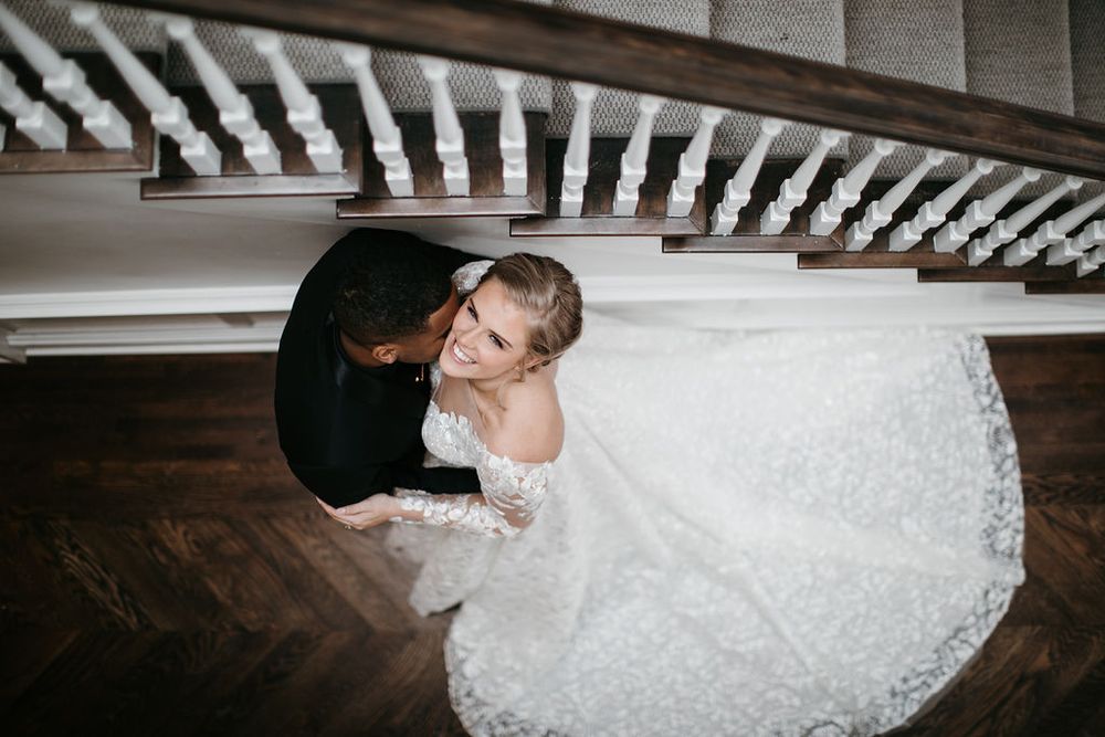 Aerial view of bride in lace gown and groom sharing a kiss beneath a white staircase.