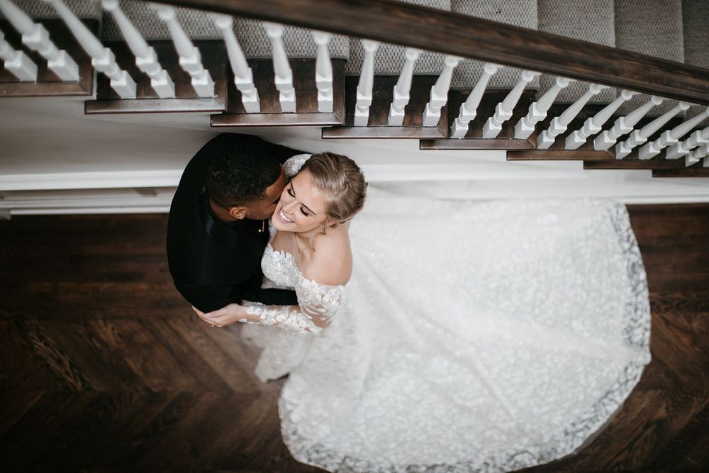 Aerial view of groom kissing bride on the cheek under a staircase, bride in elegant lace wedding gown.