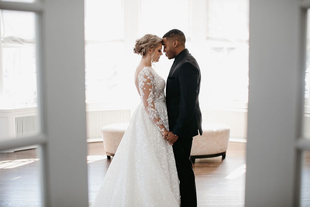 Interracial wedding couple holding hands and facing each other in a bright sunlit room with French doors.
