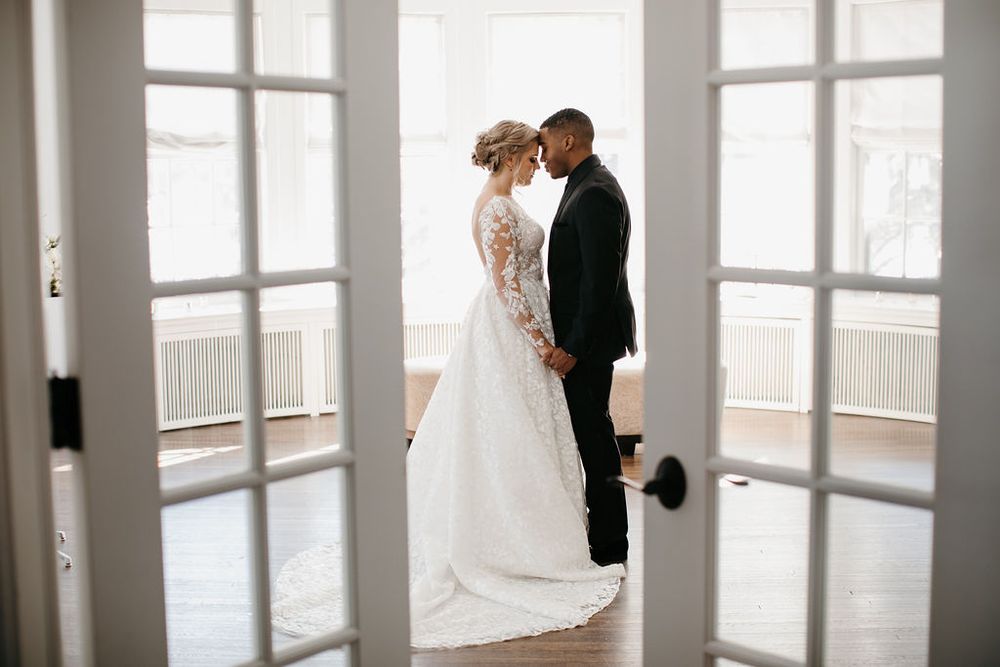 Interracial bride and groom standing face to face holding hands, photographed through open French doors.