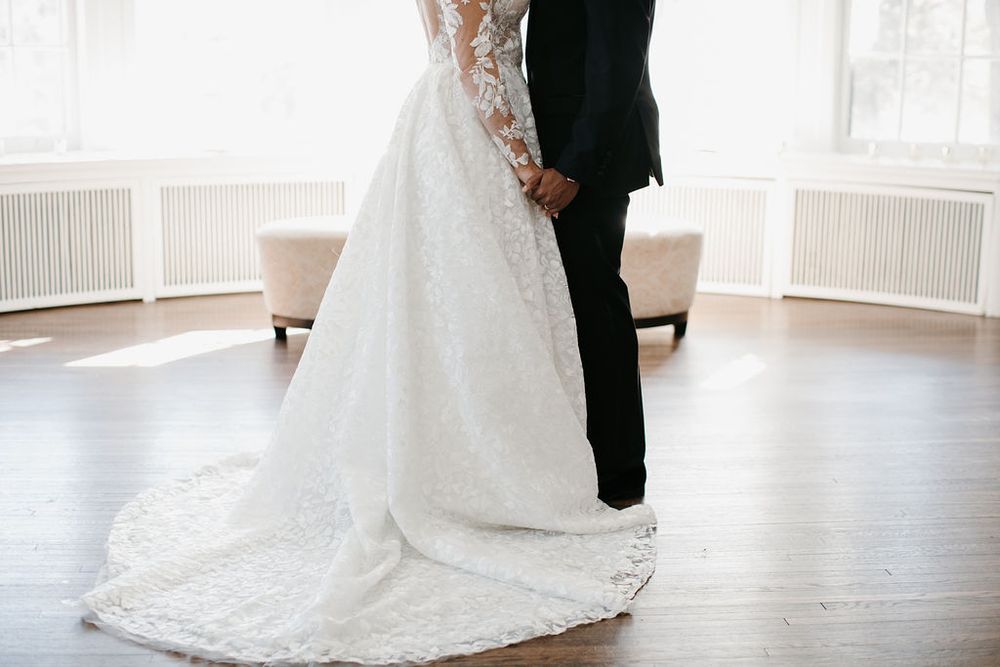 Close-up of bride and groom holding hands, showcasing intricate lace wedding dress with a long train.