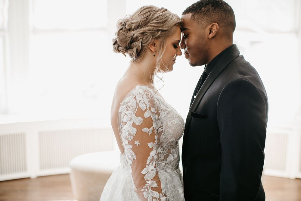 Interracial wedding couple touching foreheads romantically in a bright room, bride in long-sleeve lace gown.