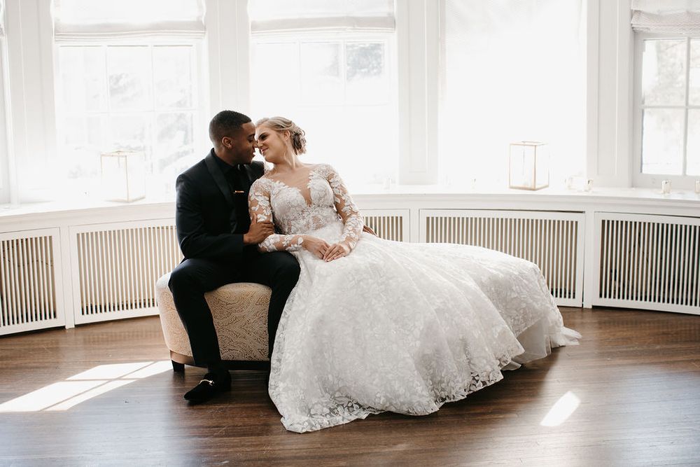 Interracial wedding couple sitting on a chaise lounge by bright windows, bride in a lace ball gown.