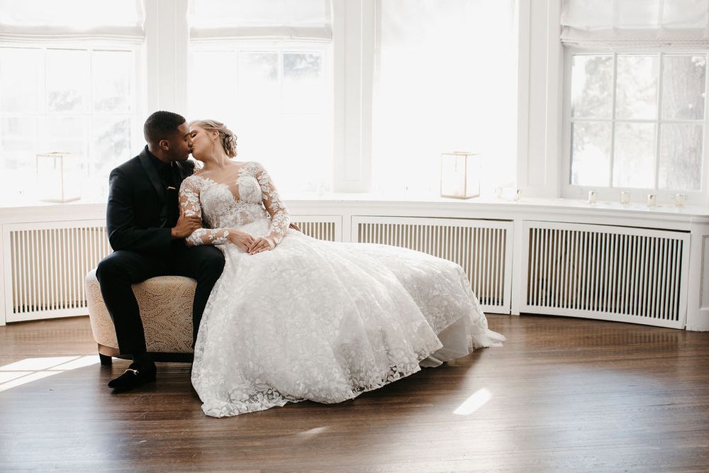 Interracial bride and groom sharing a tender kiss while seated on a chaise lounge in a sunlit room.