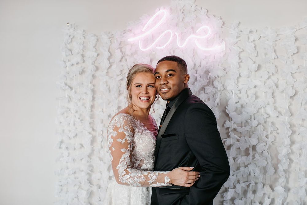 Bride and groom smiling together in front of a white floral backdrop with a pink neon love sign.