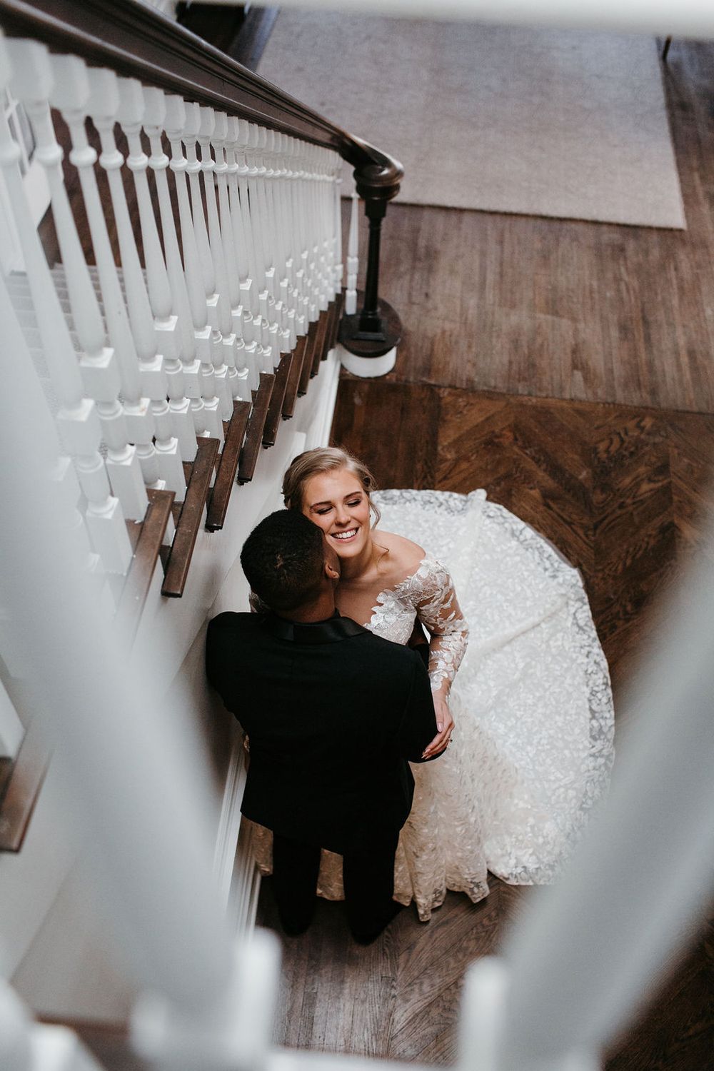 Aerial view of smiling bride in lace gown and groom embracing on wooden staircase inside elegant venue.