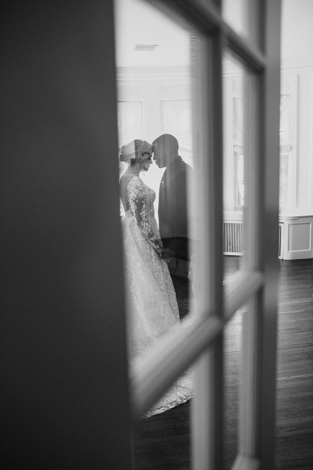 Black and white photo of bride and groom sharing intimate moment viewed through glass door panels.