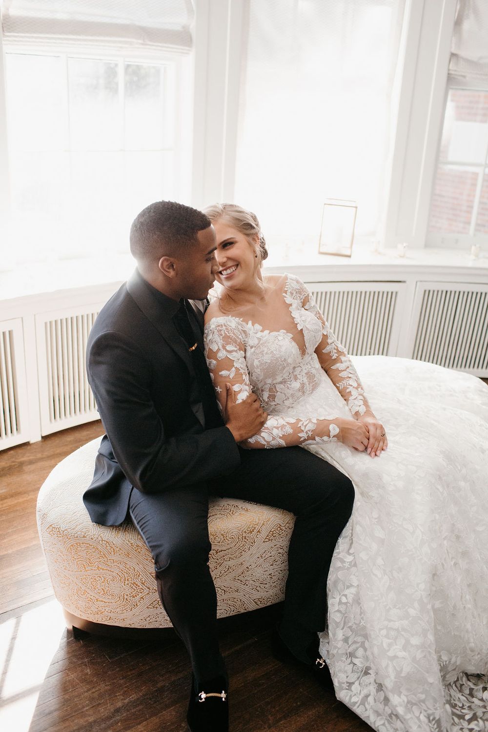 Bride in lace gown laughing with groom in black suit seated on round ottoman by windows.