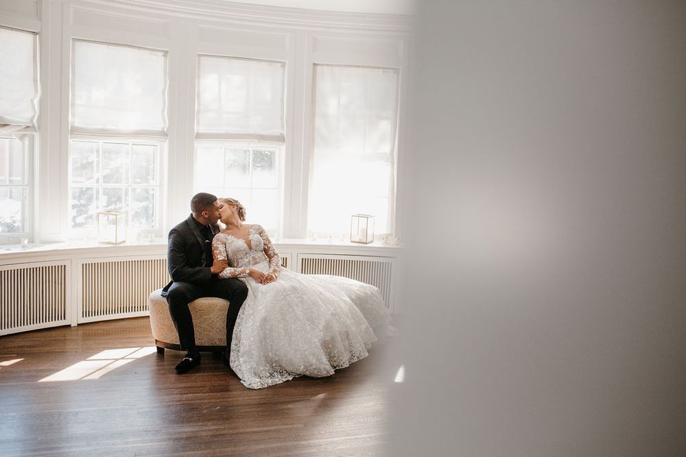 Interracial couple sharing a kiss while seated on ottoman in sunlit bay window room.