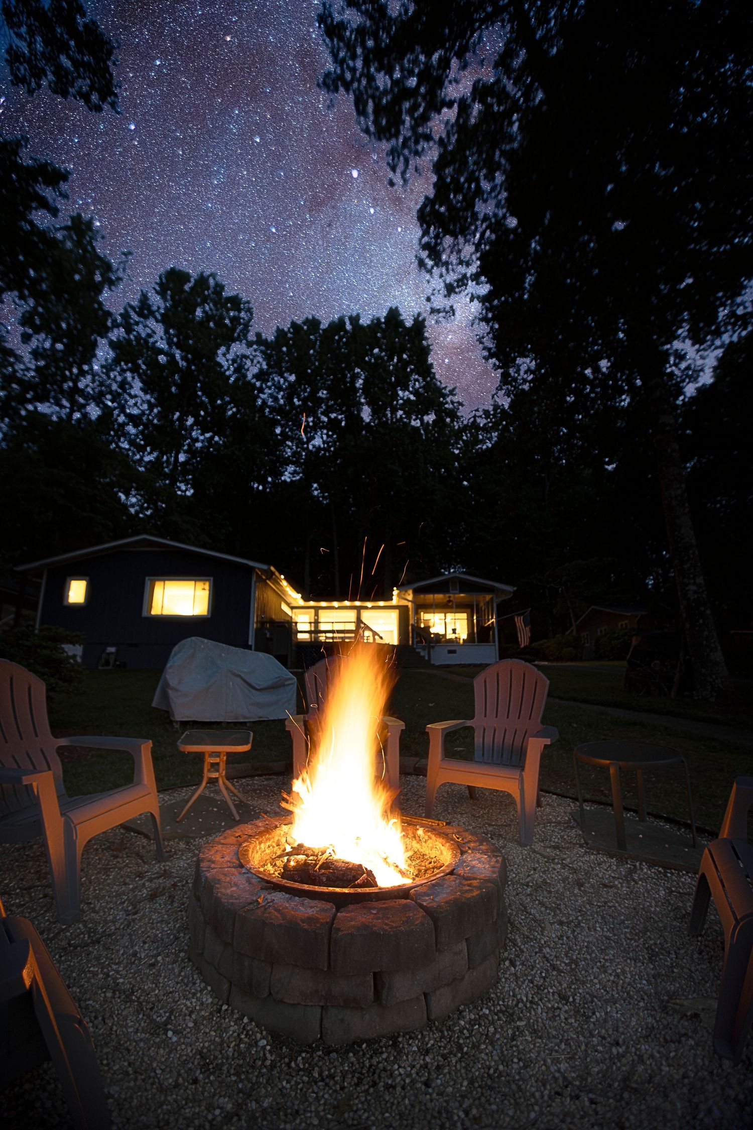 Cozy outdoor fire pit scene with Adirondack chairs illuminated under a starry night sky.