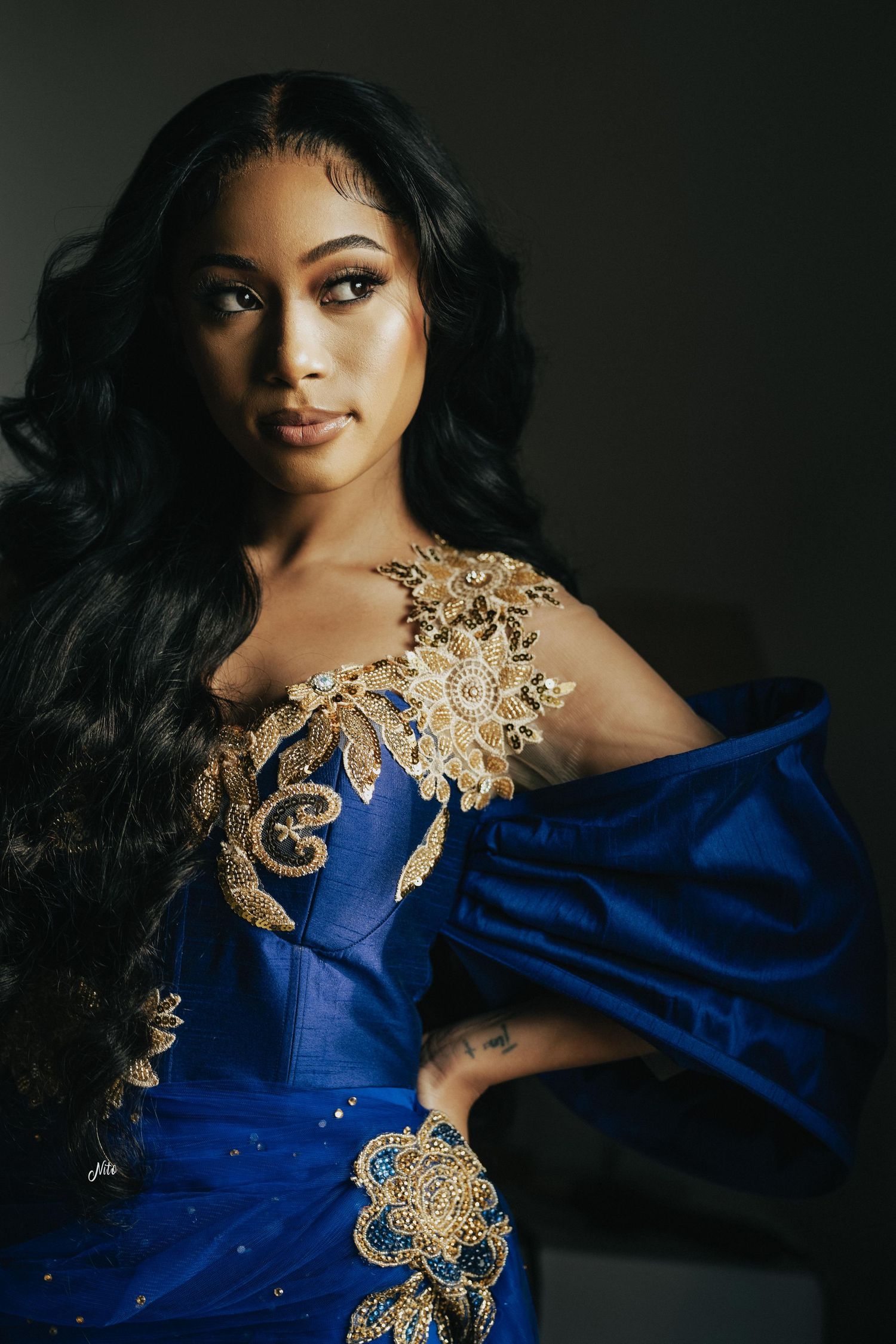 Dramatic portrait in a royal blue gown with intricate gold embellishments against dark background.