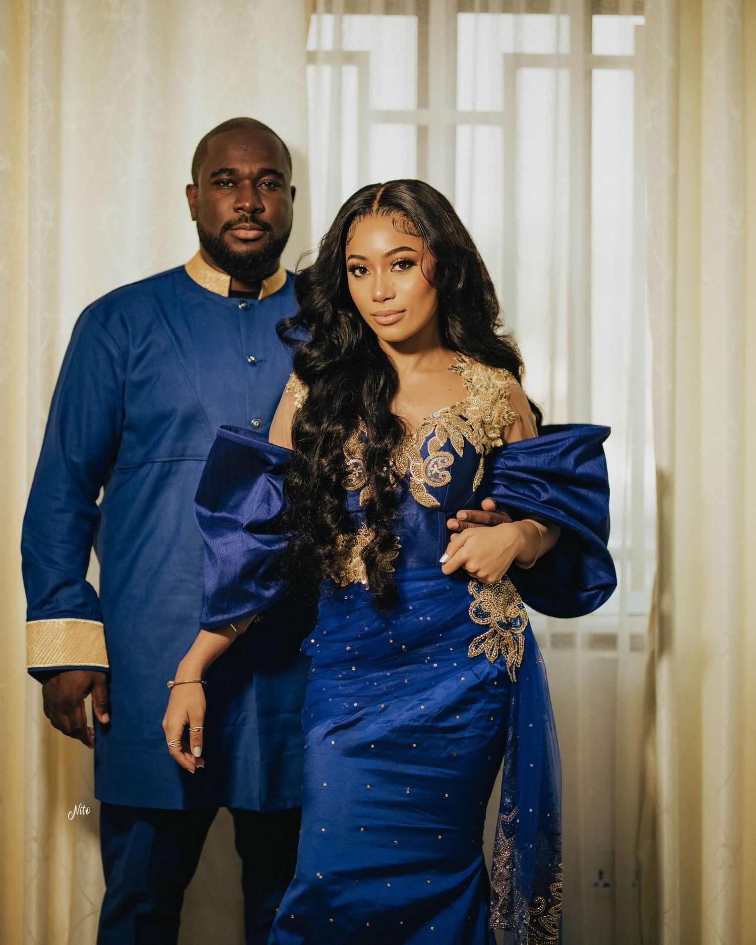 Formal portrait in coordinated blue and gold traditional African attire against light curtained background.