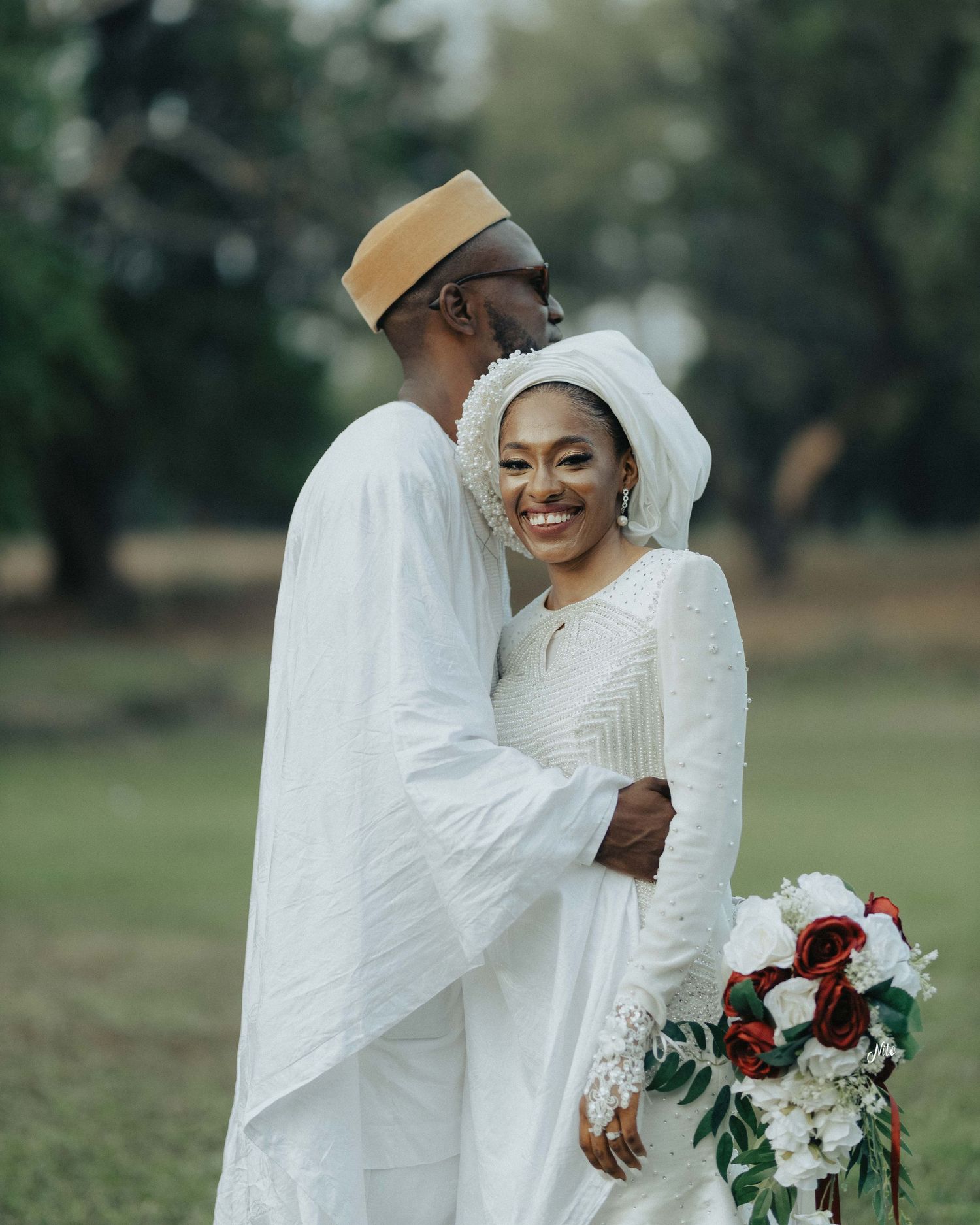 A wedding couple in white traditional attire embraces while holding a bouquet of red and white flowers.