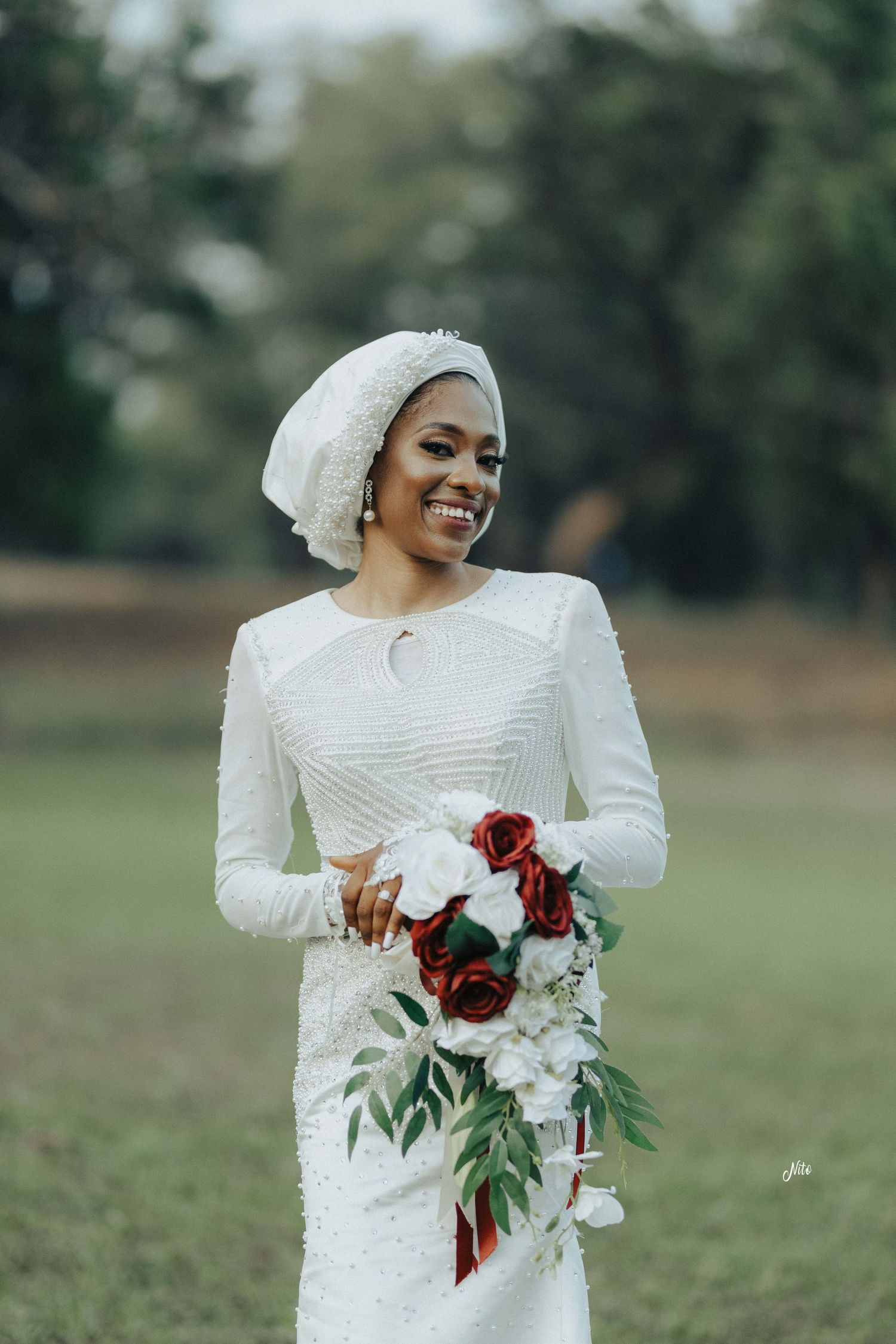 A bride in an elegant white gown holds a red and white bouquet while smiling outdoors.