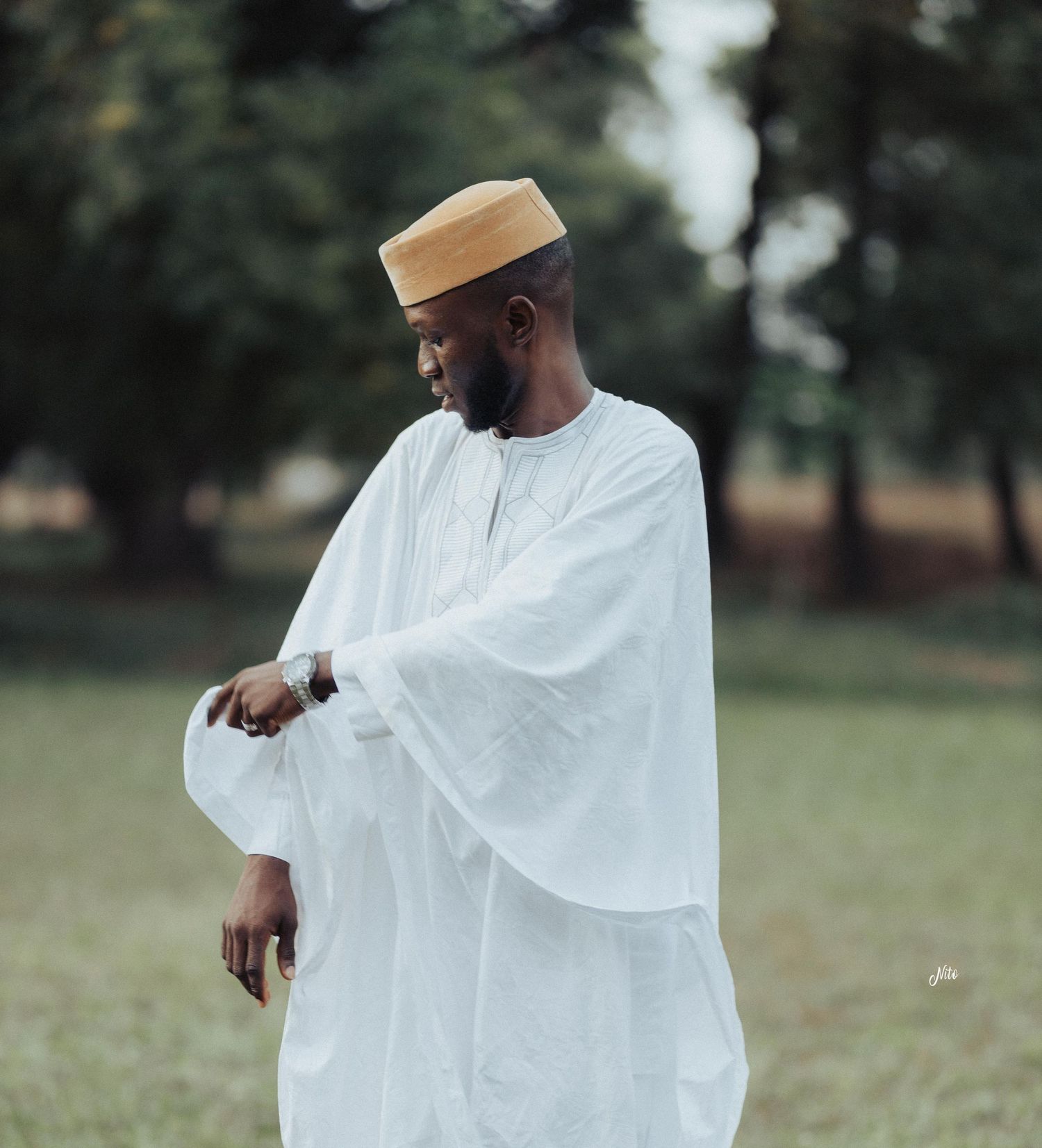 A person wearing traditional white African attire and a tan cap stands thoughtfully in an outdoor setting.