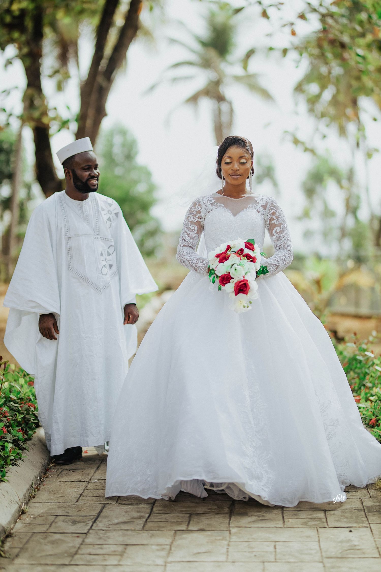 A wedding couple in traditional white attire poses on a stone path surrounded by tropical greenery.