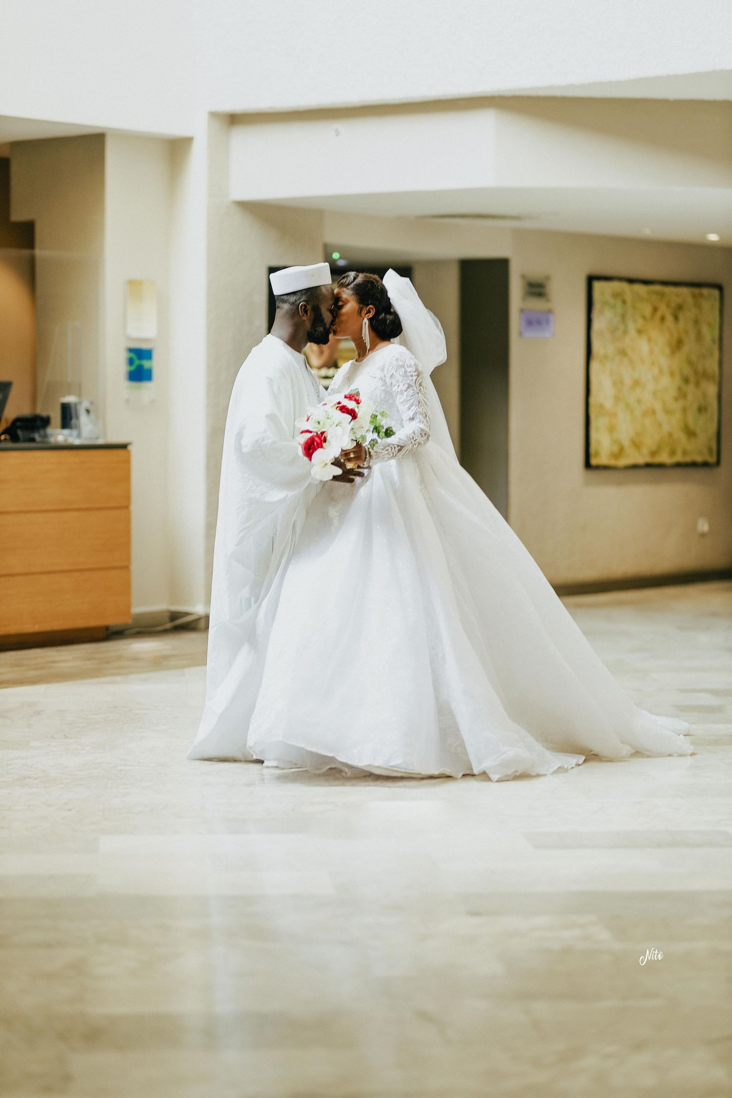 A wedding couple in matching white attire shares an intimate moment indoors.