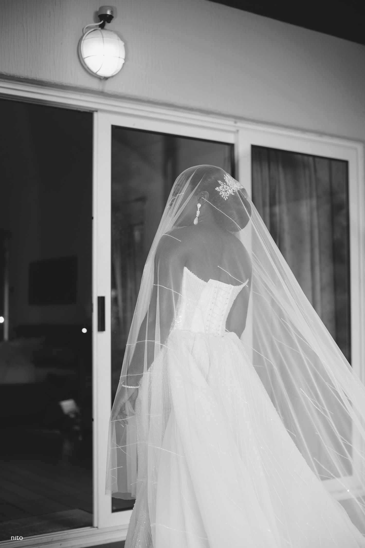 A flowing white wedding gown and veil captured in black and white against a sliding door.