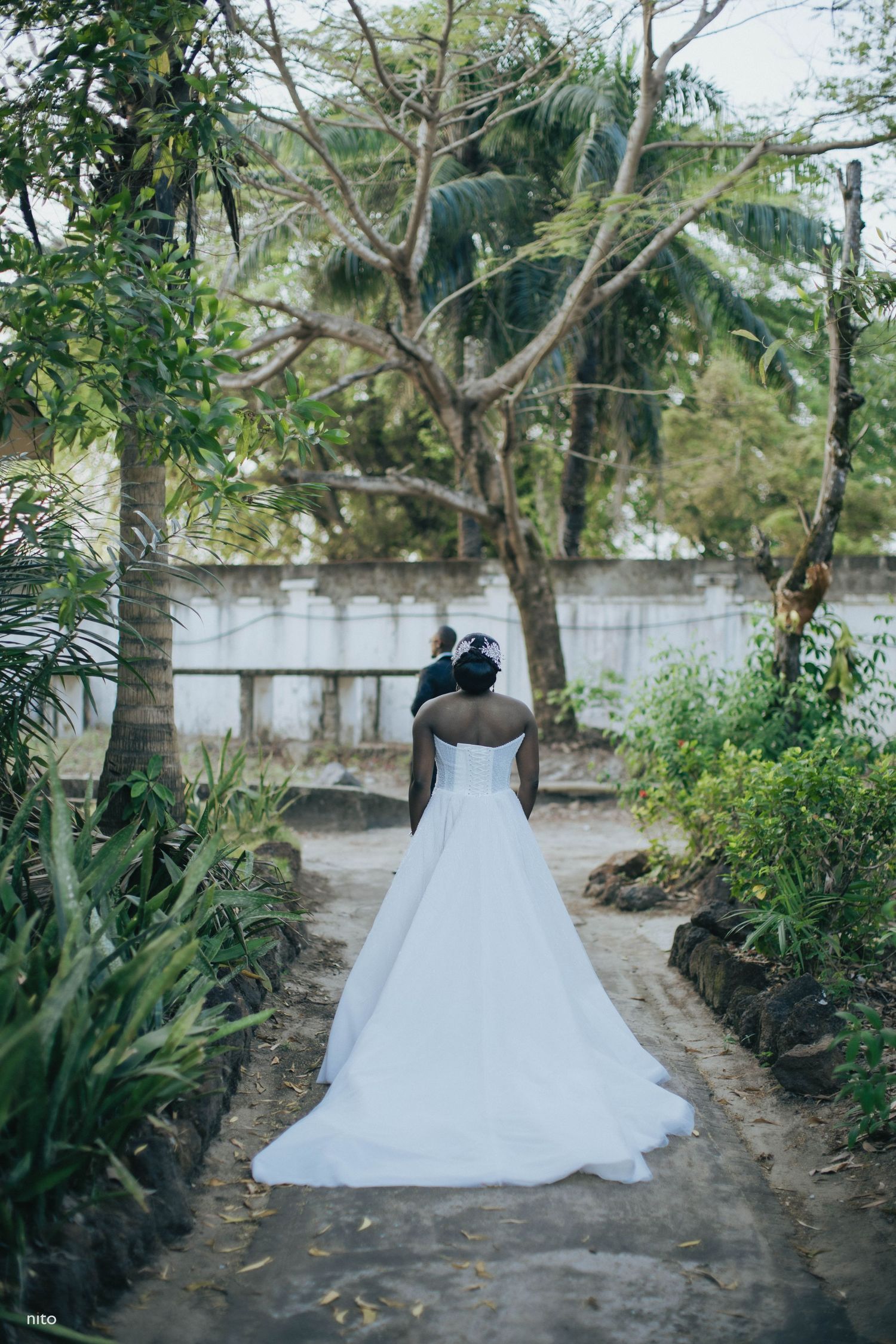A bride in a white strapless wedding dress stands on a garden pathway surrounded by tropical plants.