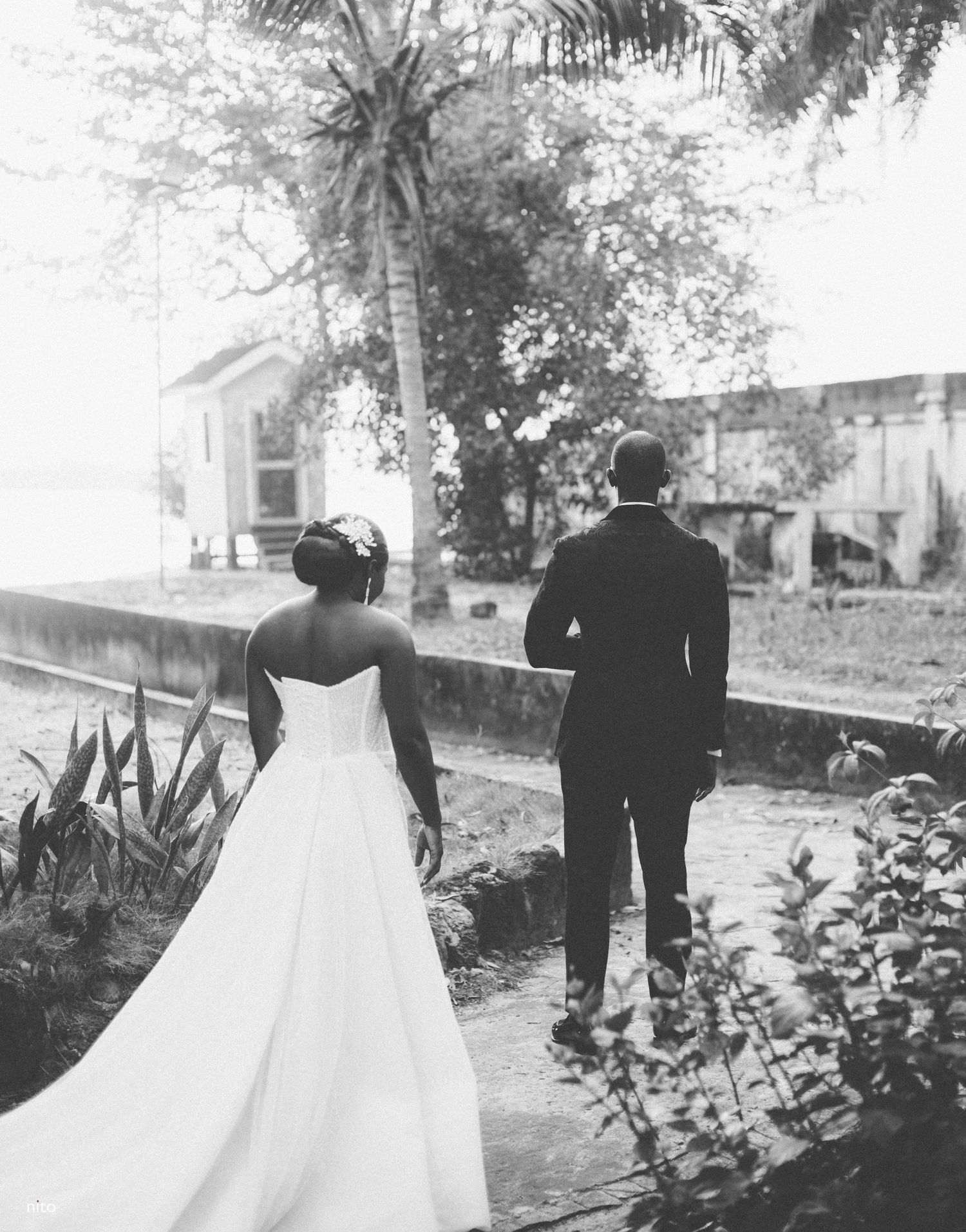 A couple in wedding attire walks along a tree-lined path in black and white.
