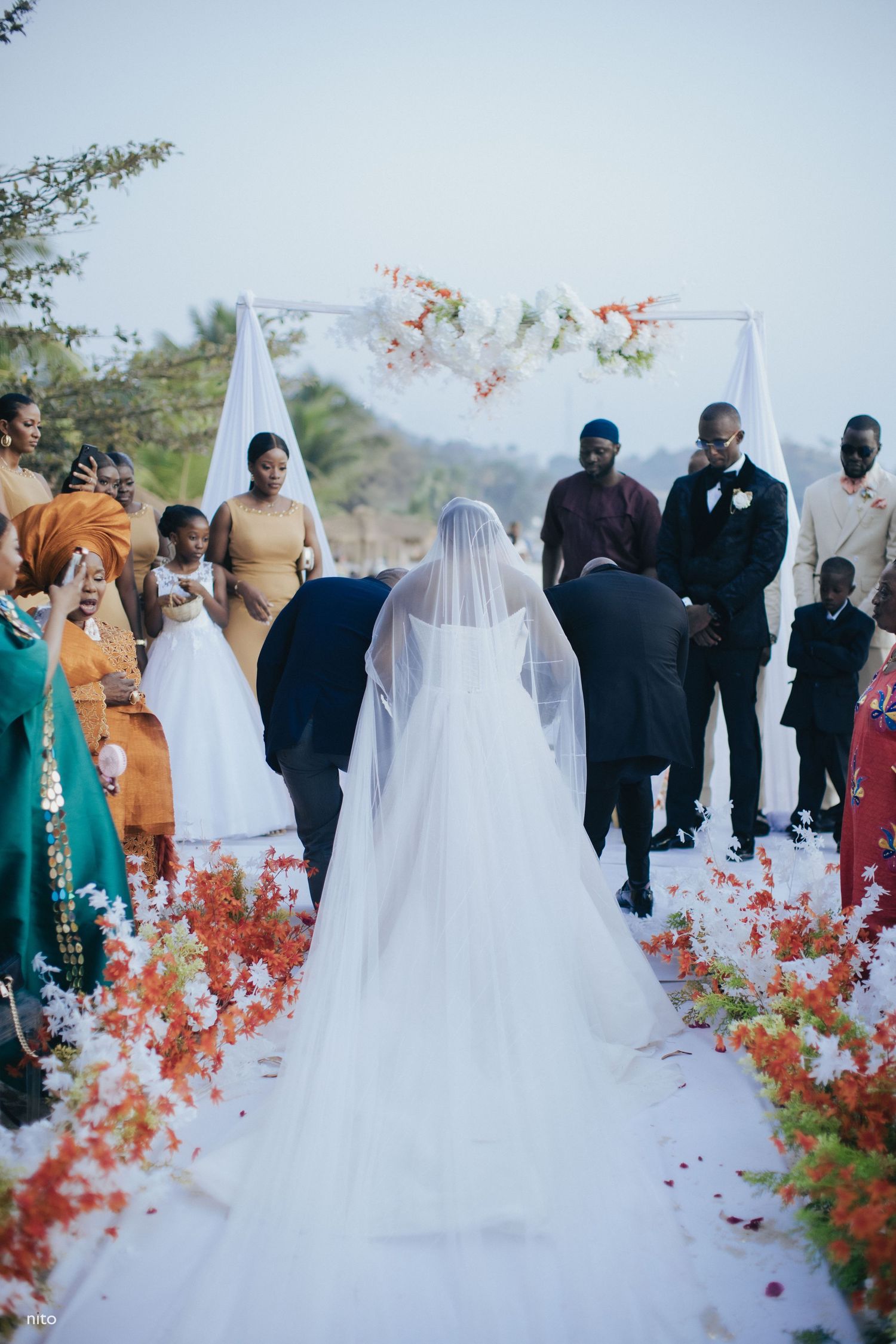 A bride walking down an outdoor aisle decorated with coral flowers towards a floral arch.