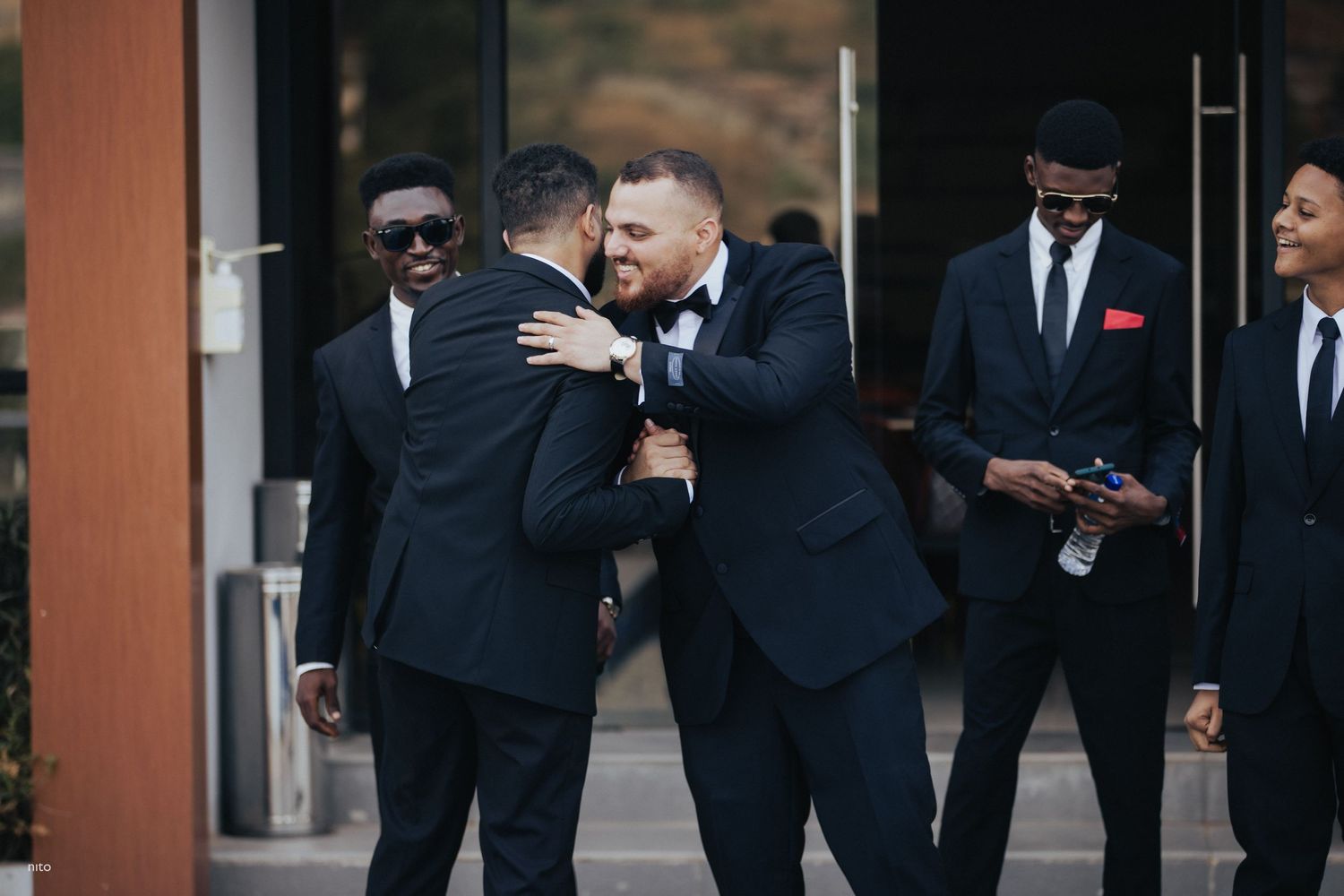 Group of groomsmen in formal suits sharing emotional moment outside venue.