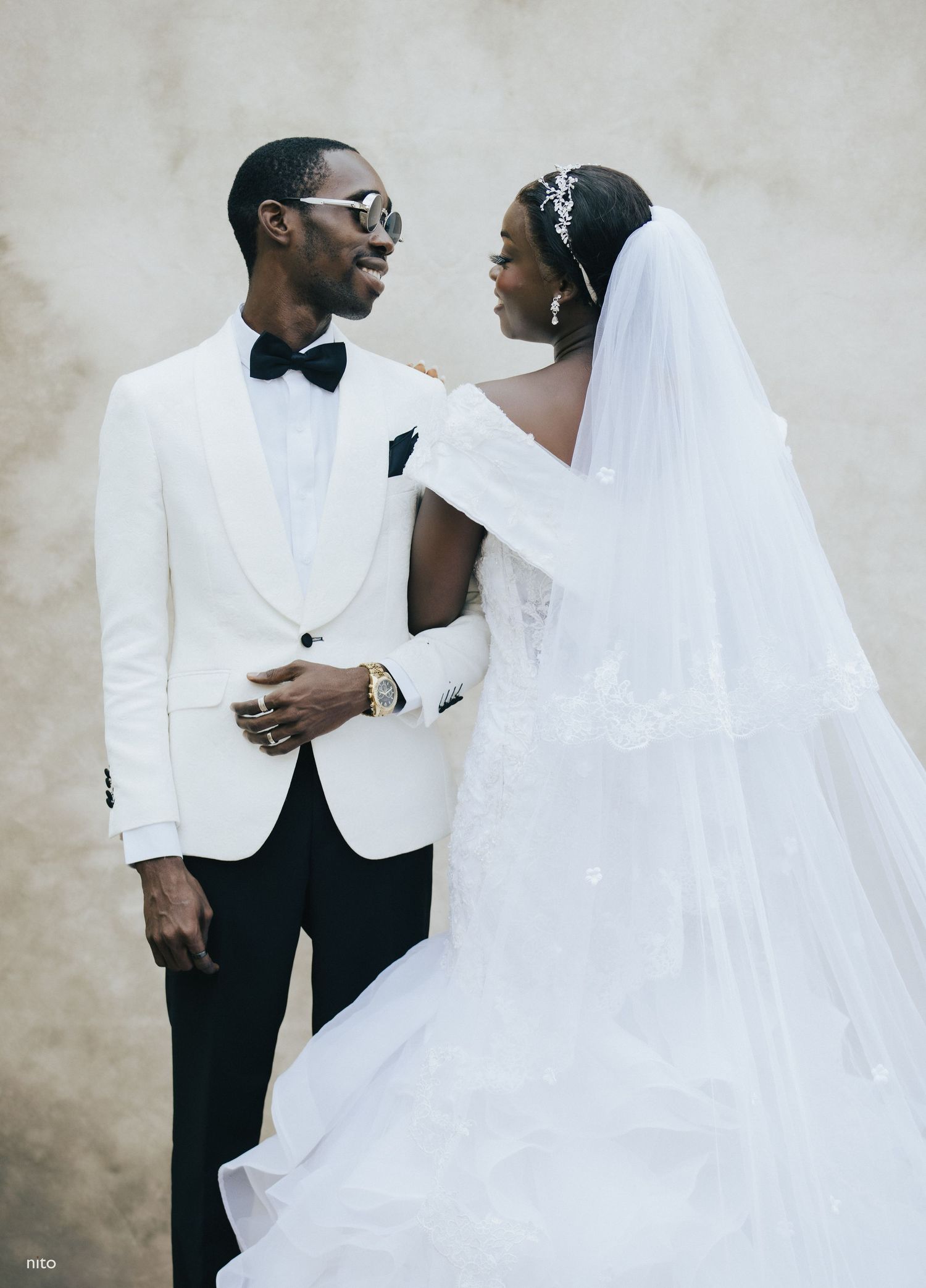 Stylish wedding portrait featuring white tuxedo jacket and flowing bridal veil against neutral backdrop.