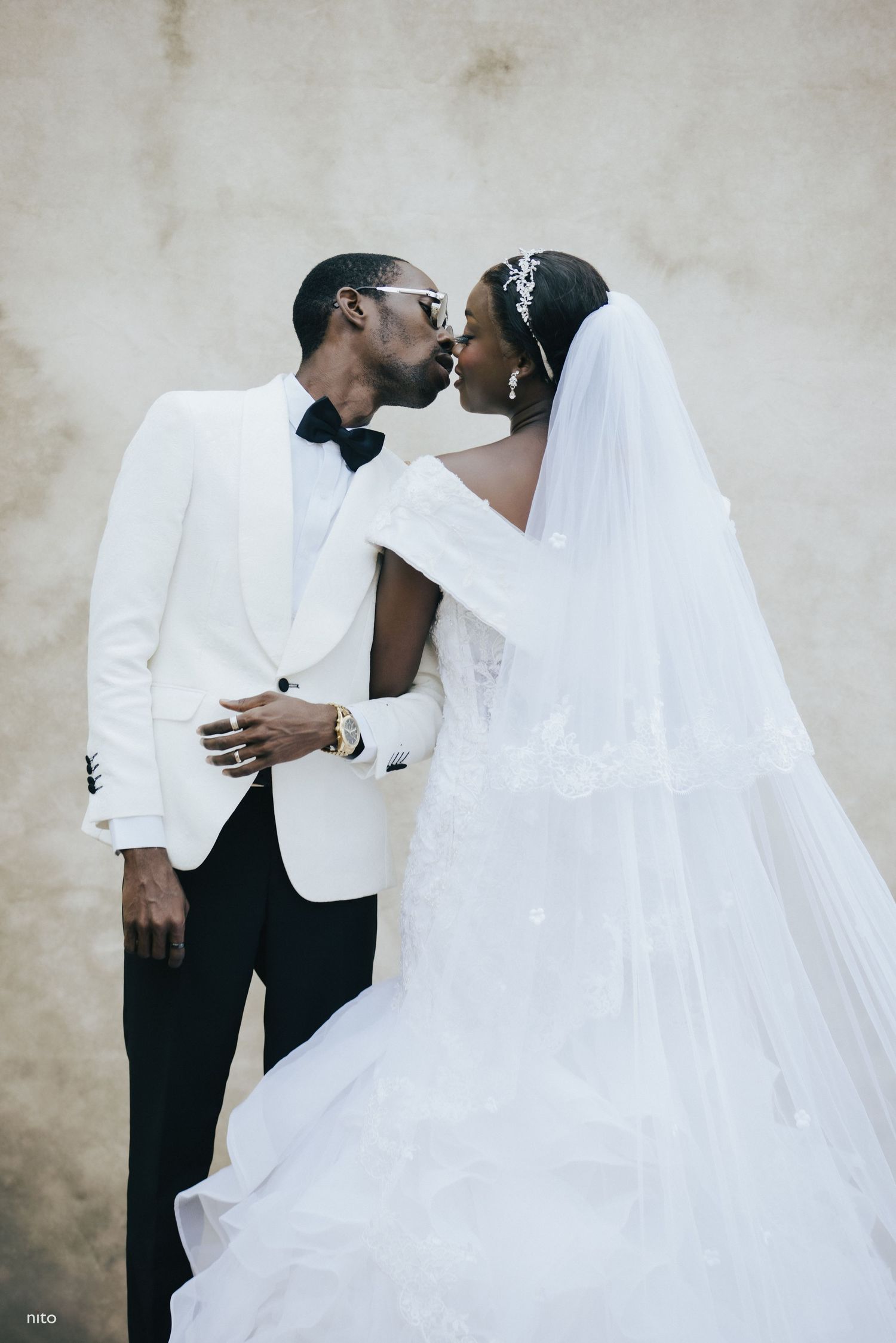 Romantic wedding portrait featuring classic white tuxedo and flowing bridal gown against neutral wall.