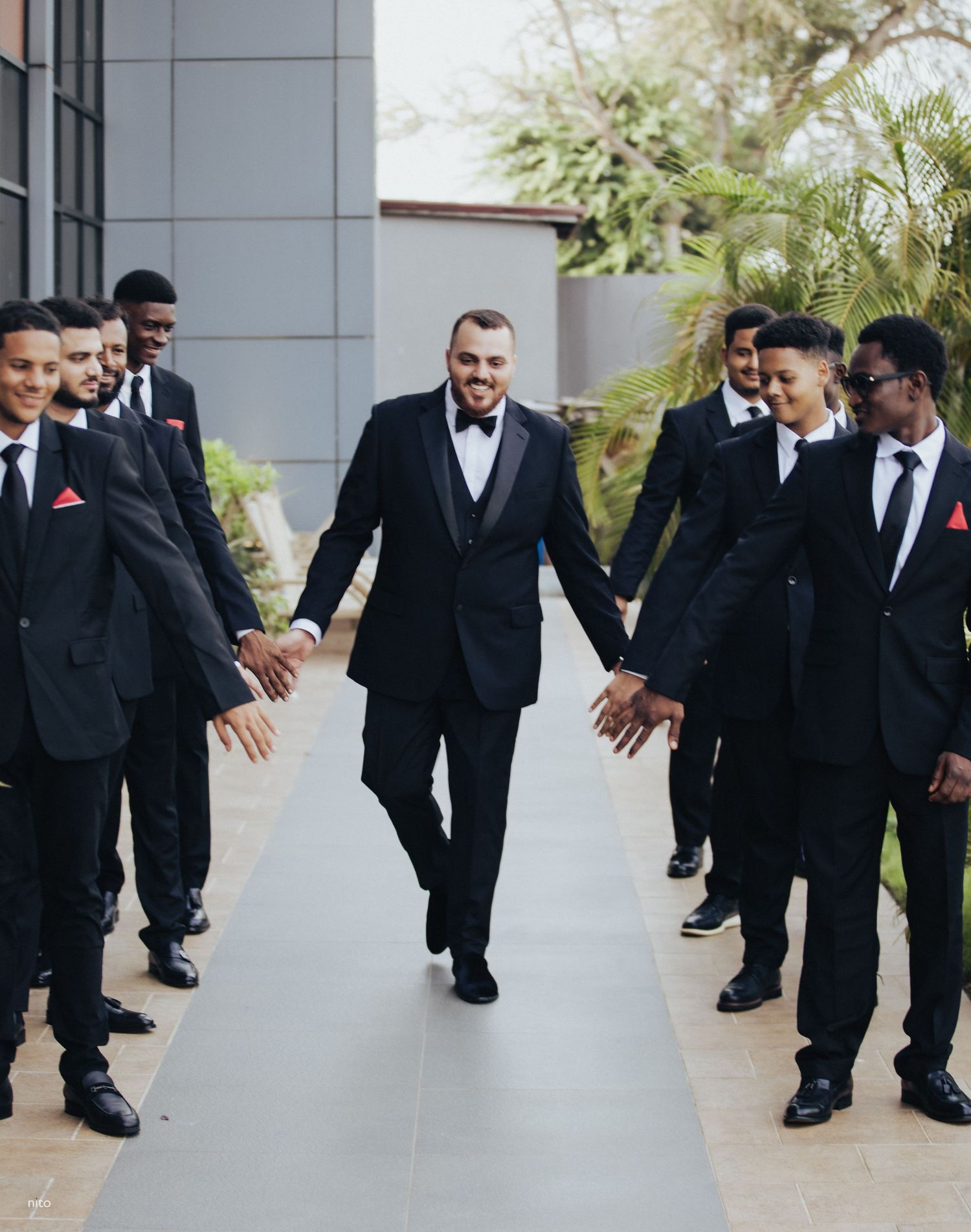 Wedding party in black suits walking confidently down outdoor pathway.