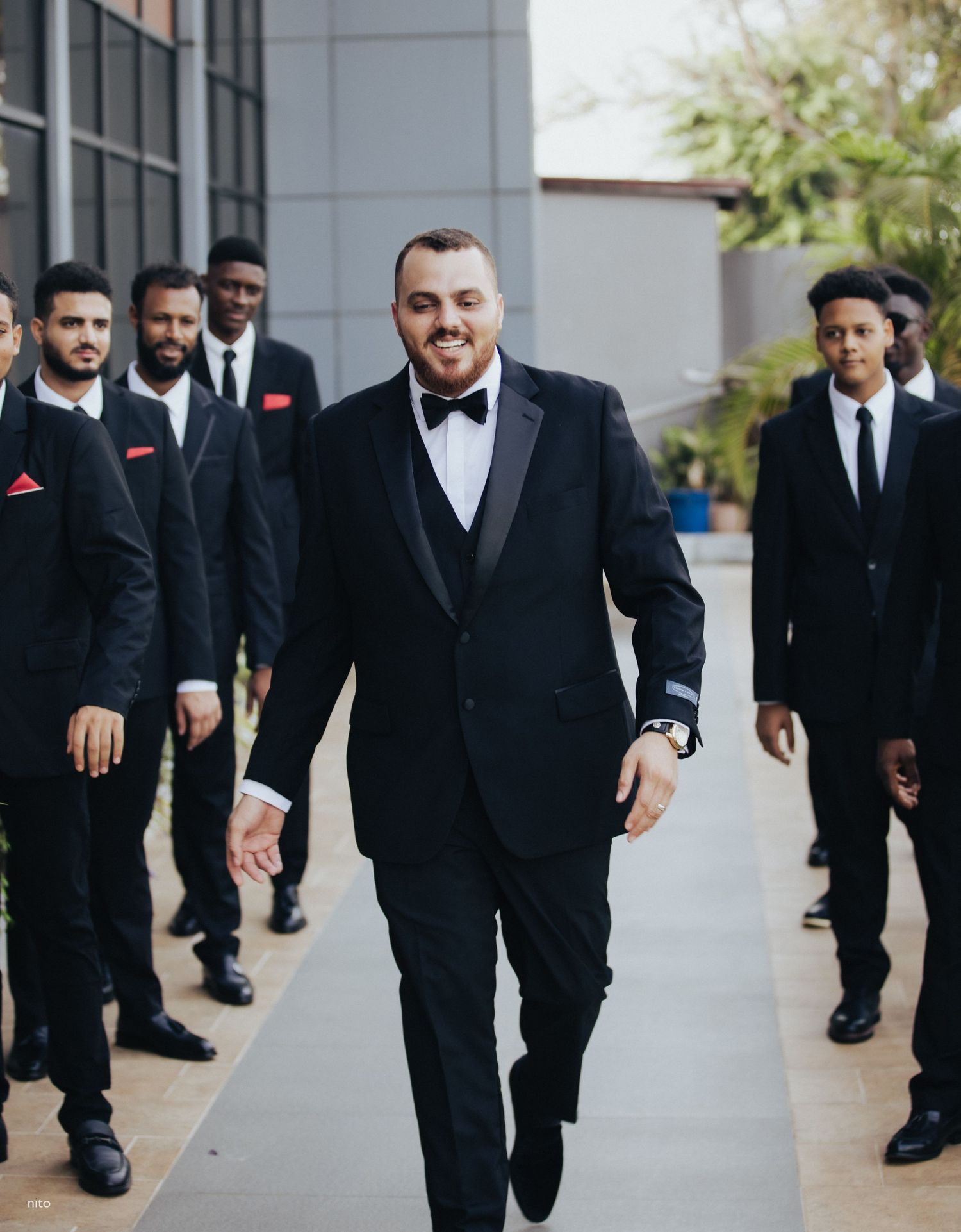 Stylish groomsmen in black tuxedos walking together outside modern building.