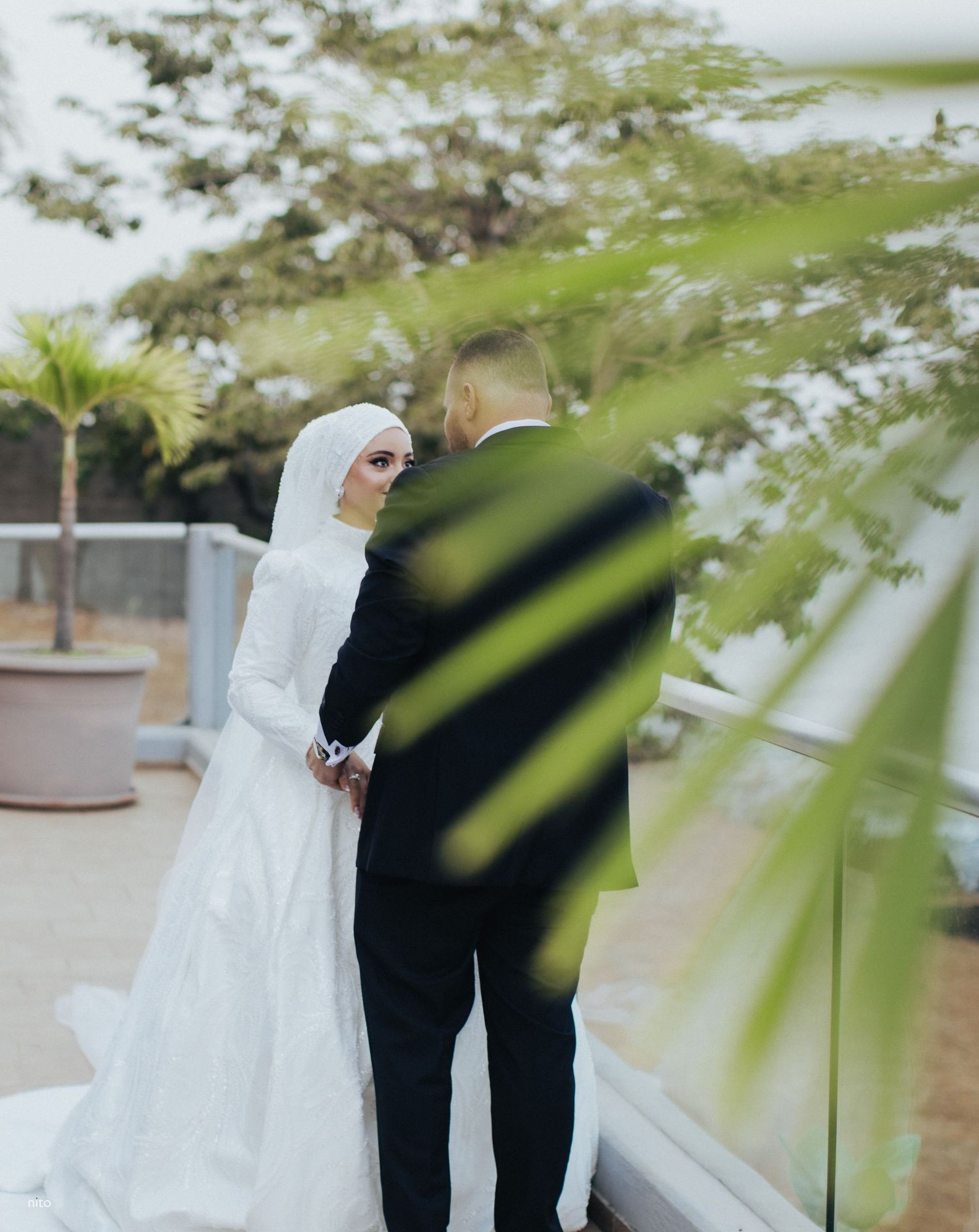 A couple shares an intimate moment during their wedding photoshoot among palm trees and greenery.