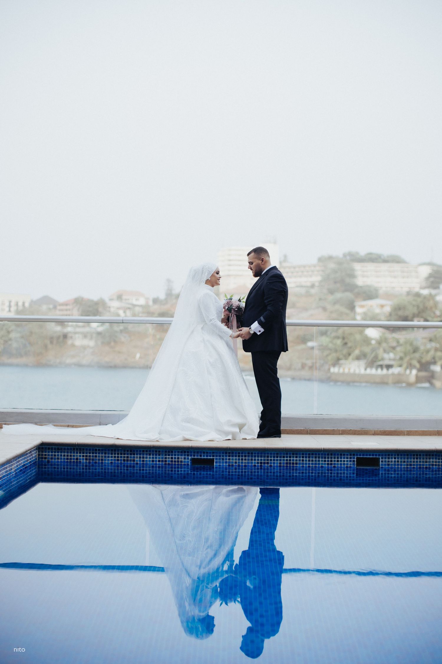 A wedding couple stands beside a pool with their reflection visible in the blue water.