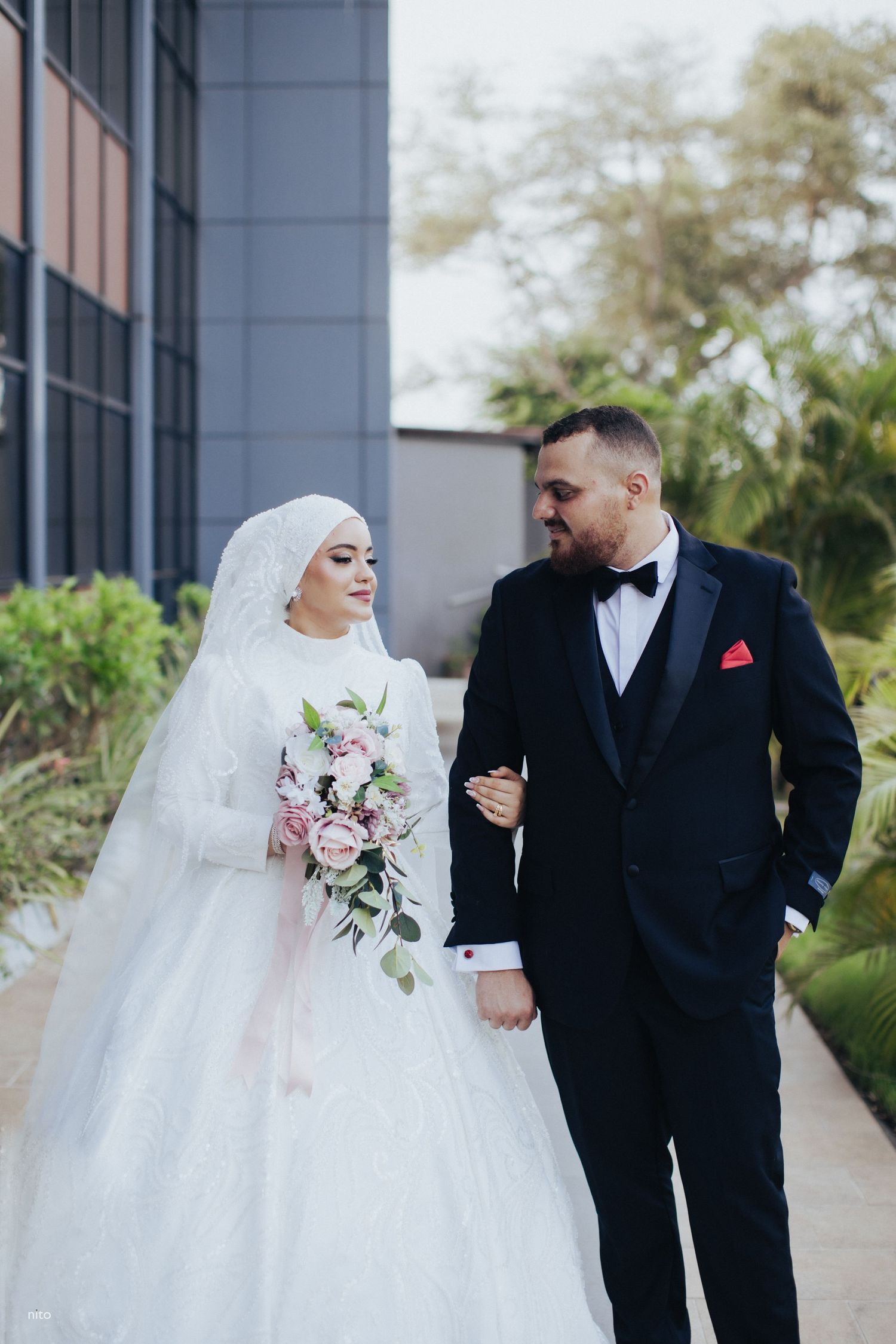 A couple poses for a modern wedding photo with pink bouquet against a contemporary building backdrop.