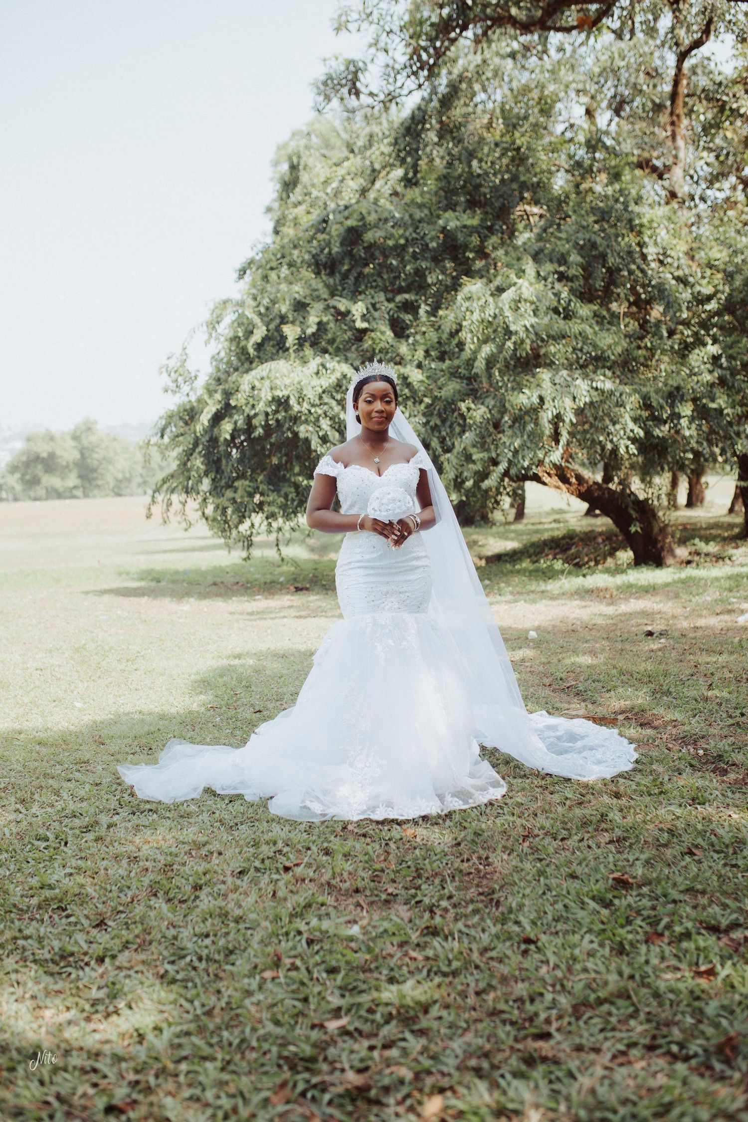 A bride in a mermaid-style wedding gown stands gracefully in a serene outdoor garden setting.