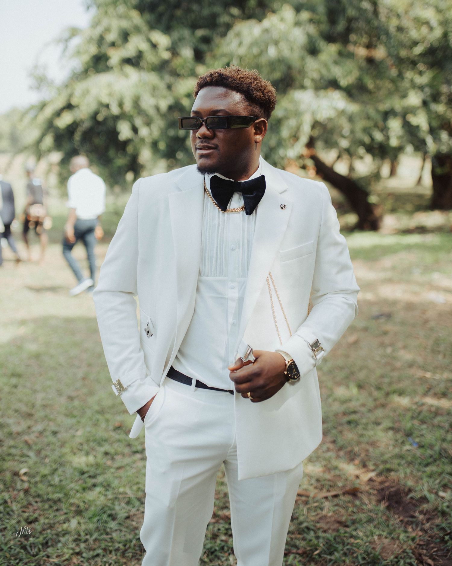 Stylish formal attire featuring a crisp white tuxedo with black bow tie and sunglasses in an outdoor garden setting.