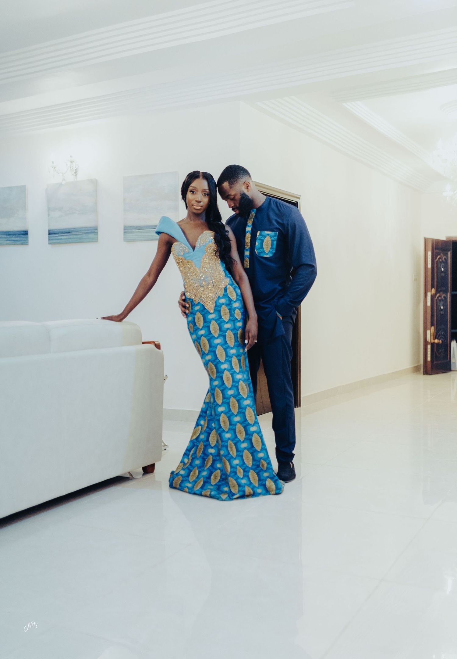 Elegant couple wearing coordinated blue and gold African-inspired formal attire in a modern white gallery space.