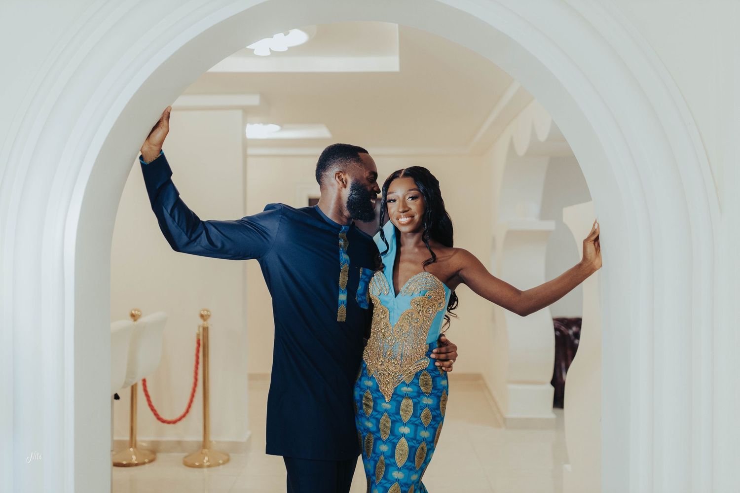 Joyful couple posing in coordinated blue African-inspired outfits against white wall background.
