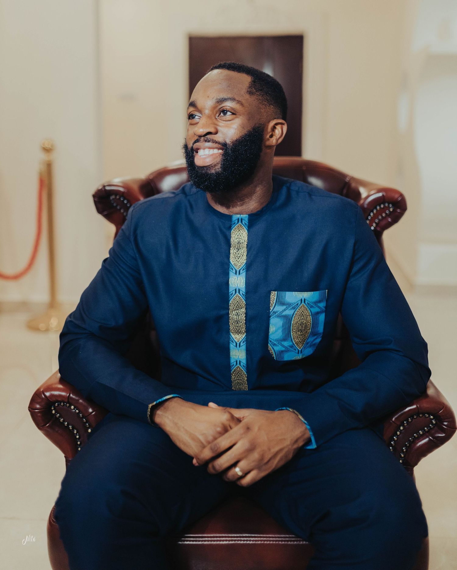 Deep blue traditional African attire with patterned details shown while seated in a leather armchair.