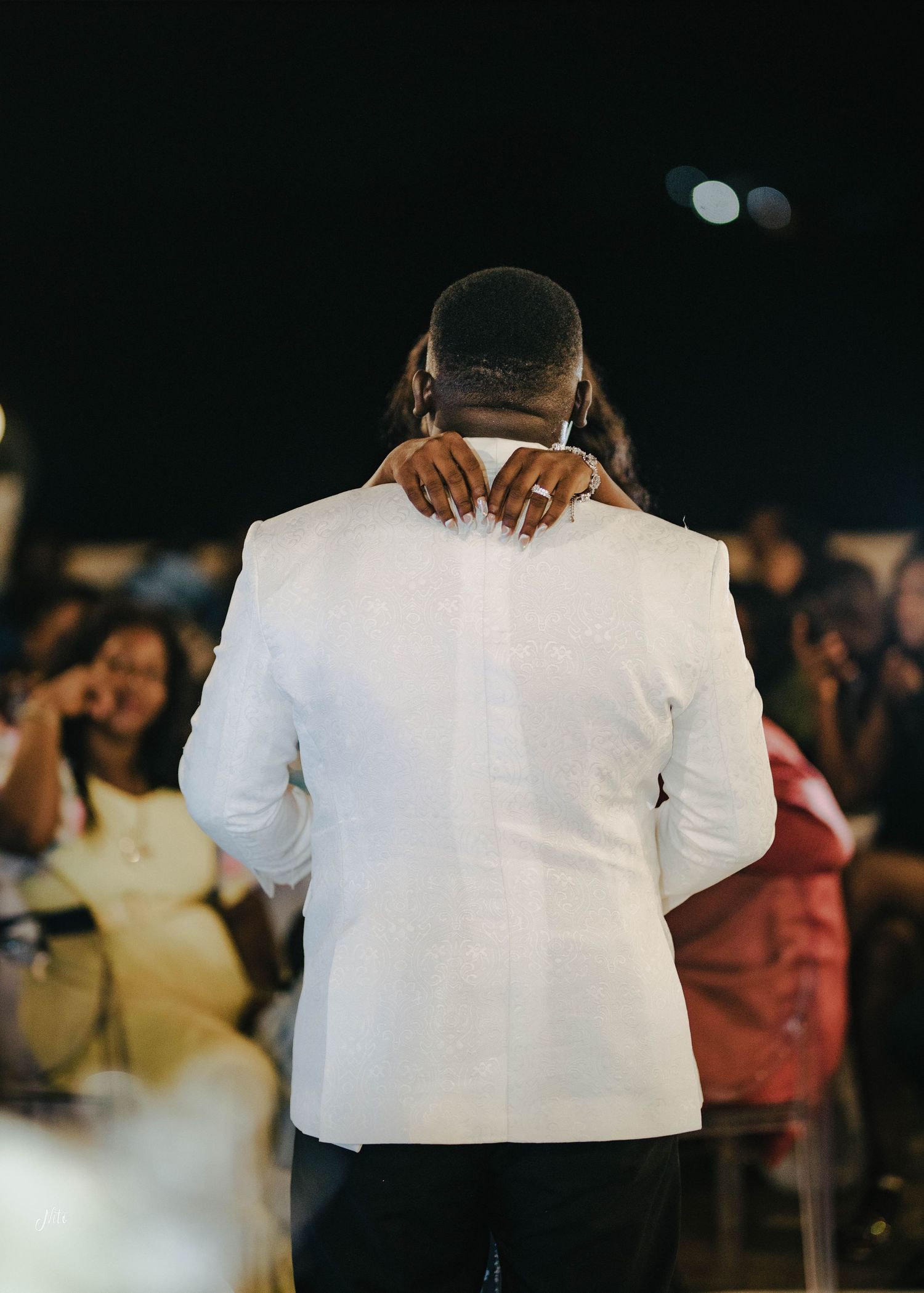 Wedding guest embracing someone in a white suit jacket during an evening reception.