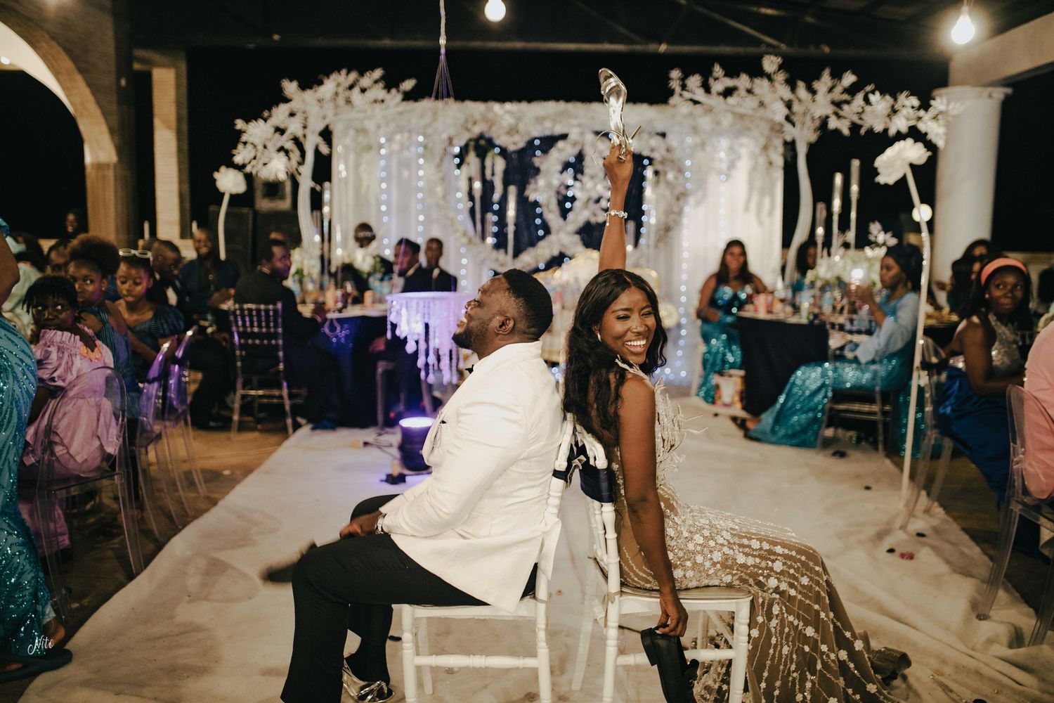 A joyful couple plays the wedding shoe game under an elegant white floral arch at their reception celebration.