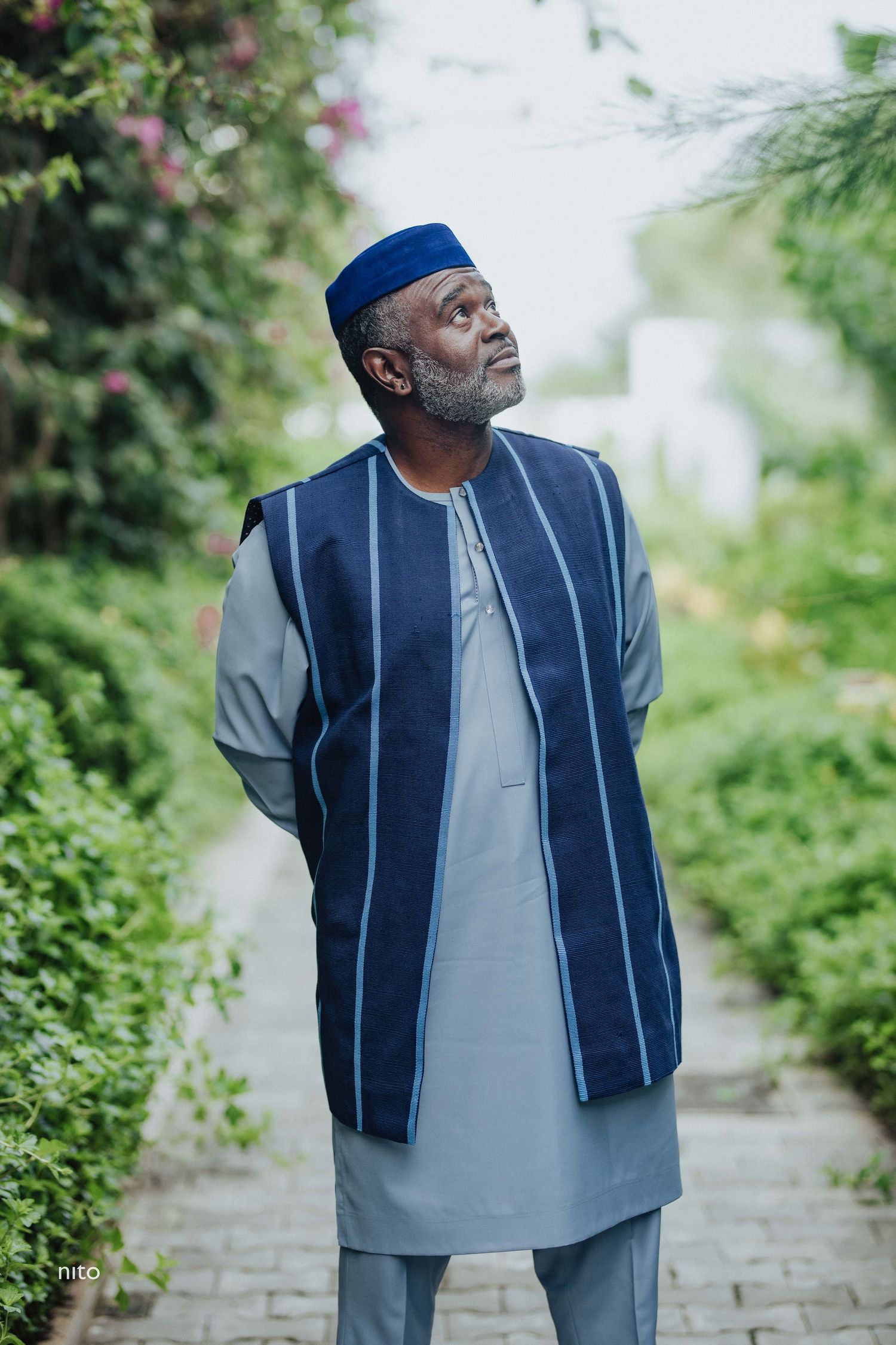 A person wearing a blue cap and traditional navy and grey clothing stands on a garden path.