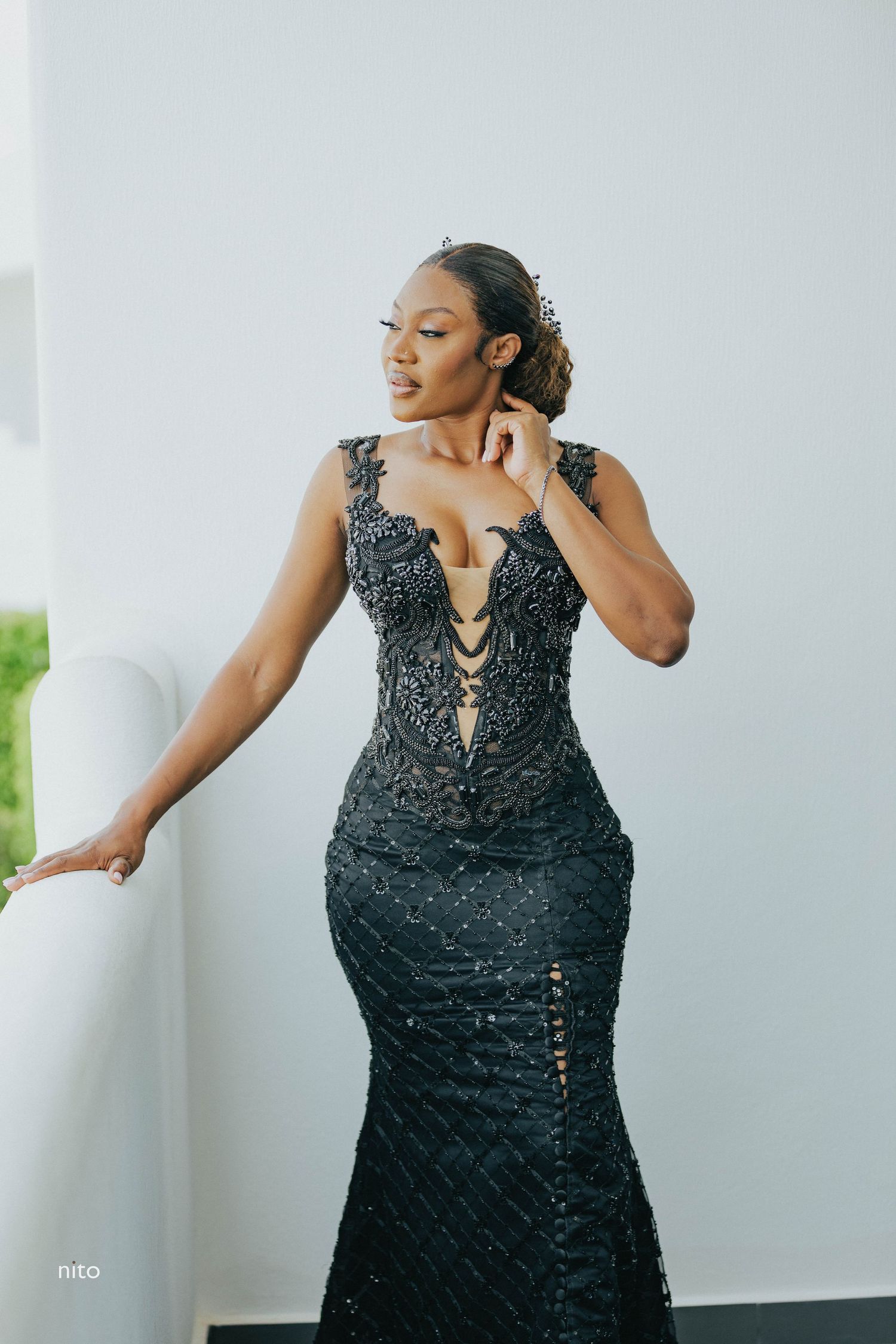 Glamorous black beaded mermaid gown with plunging neckline worn against white wall.