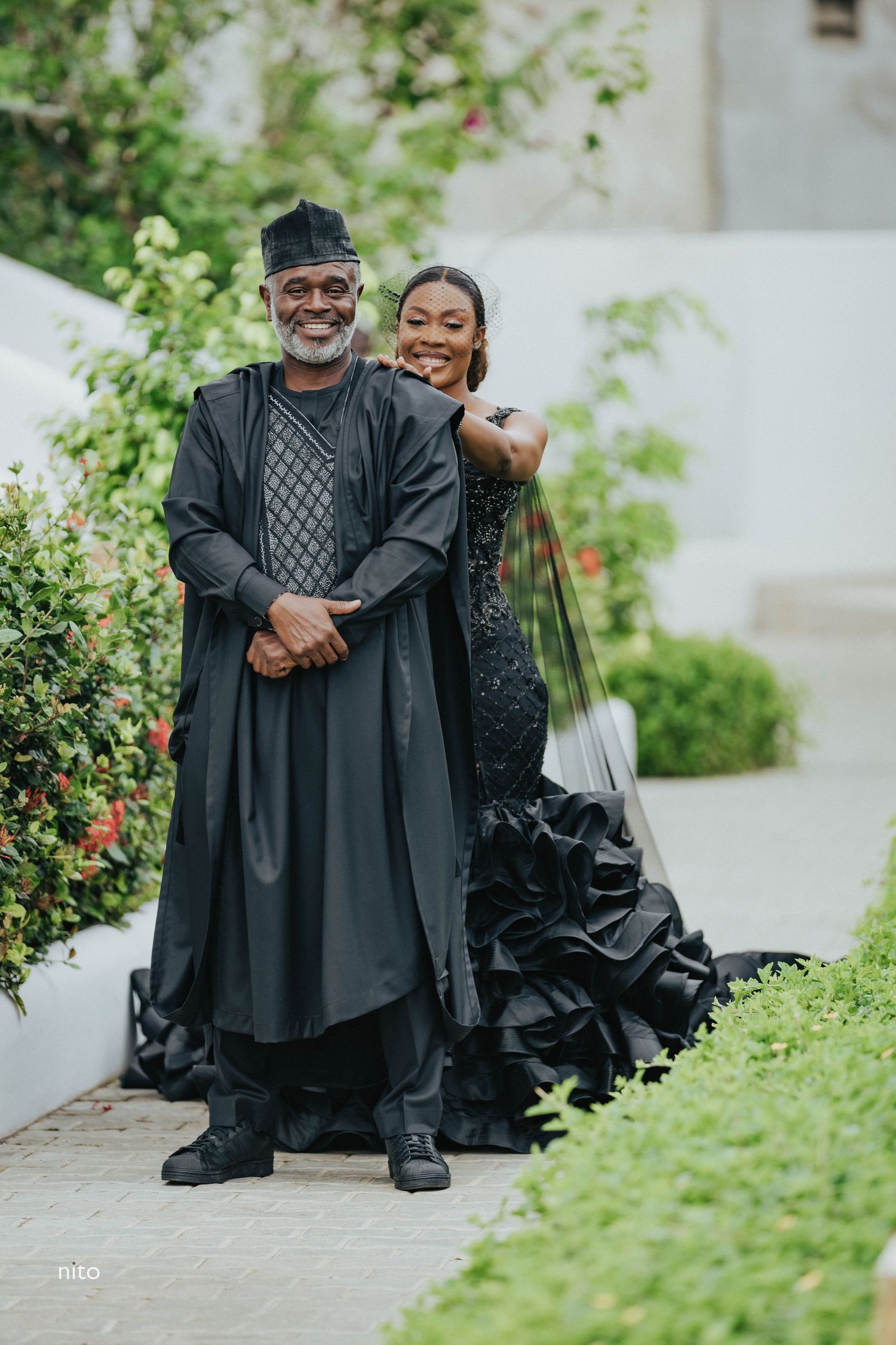 Two people in formal black attire pose together on a garden path.