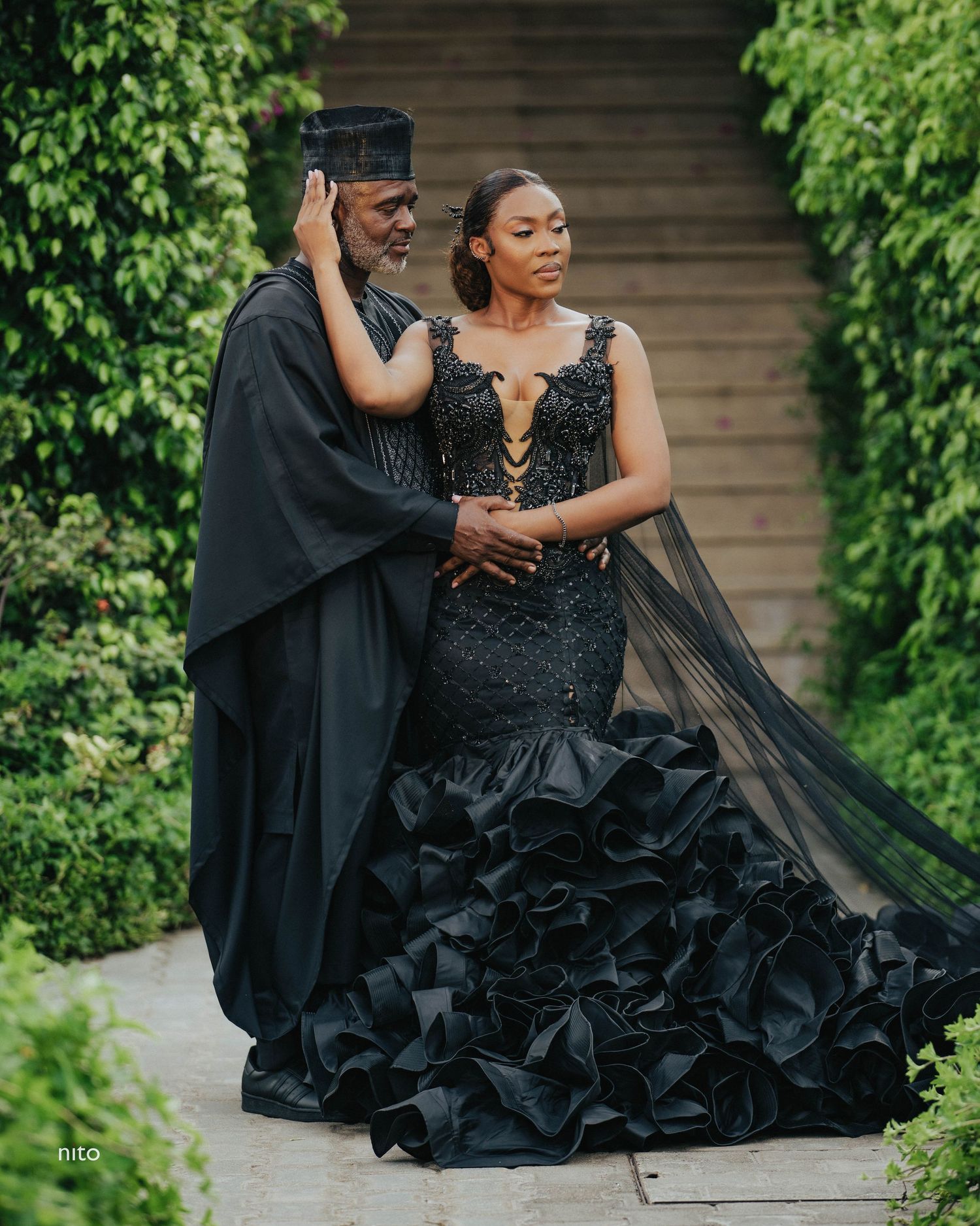 Two people in elegant black formal wear pose on garden steps.
