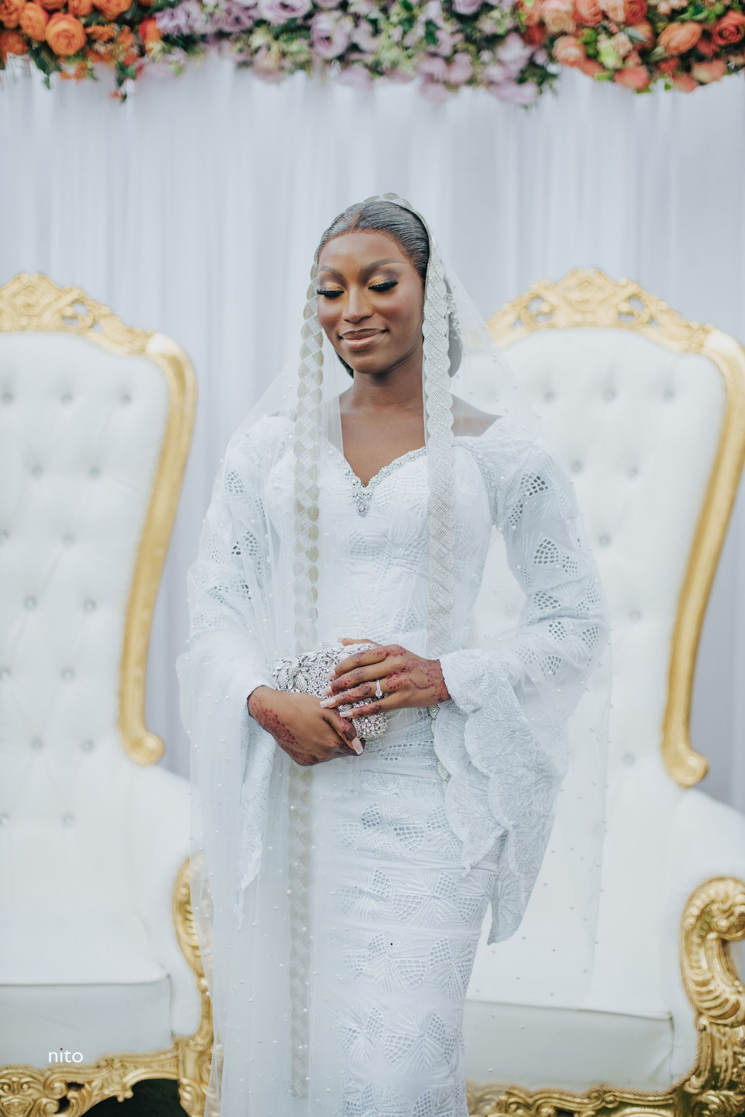 A bride in traditional white lace dress poses elegantly against a floral backdrop with gold chairs.
